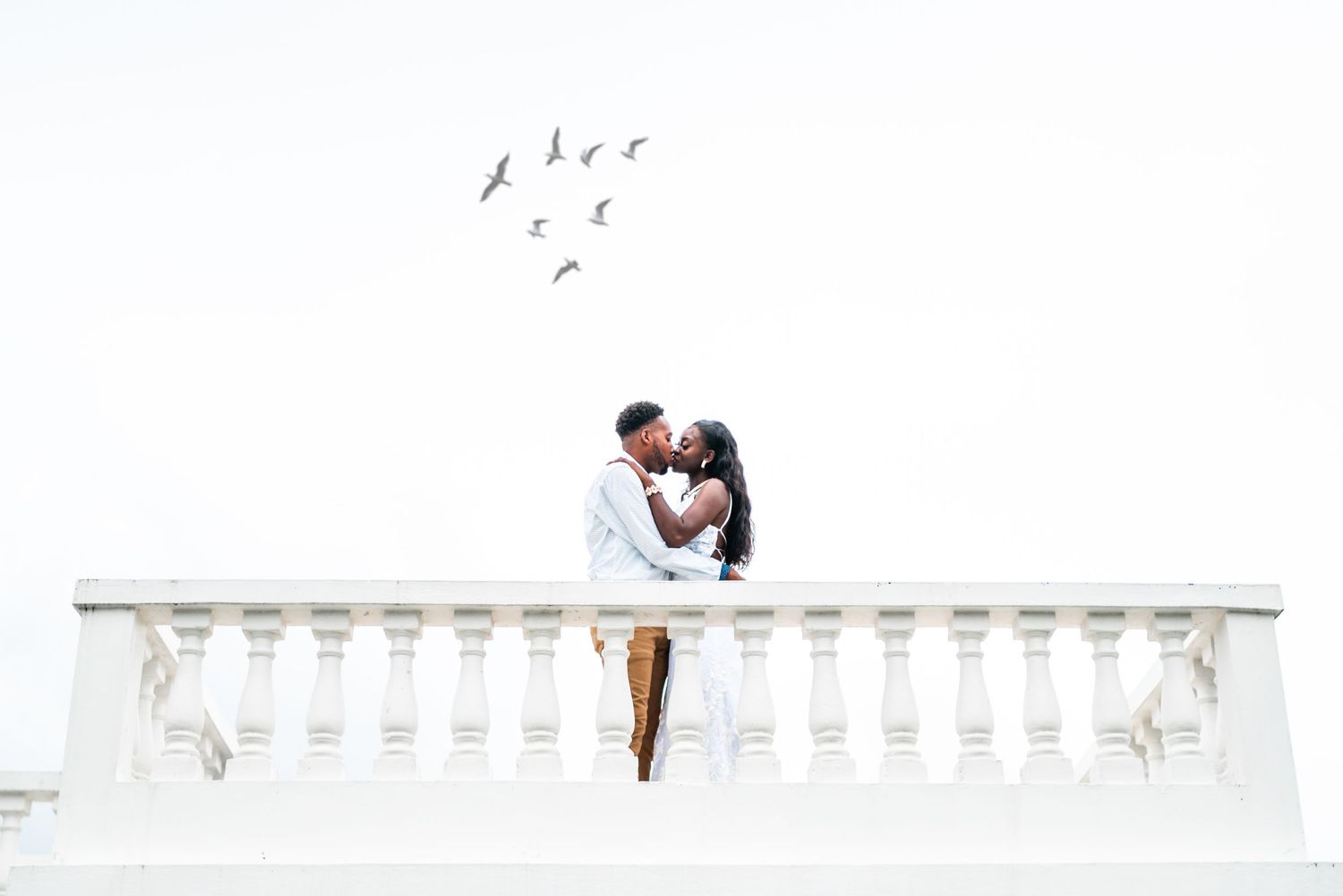 A romantic moment captured on a white balustrade with birds flying overhead.