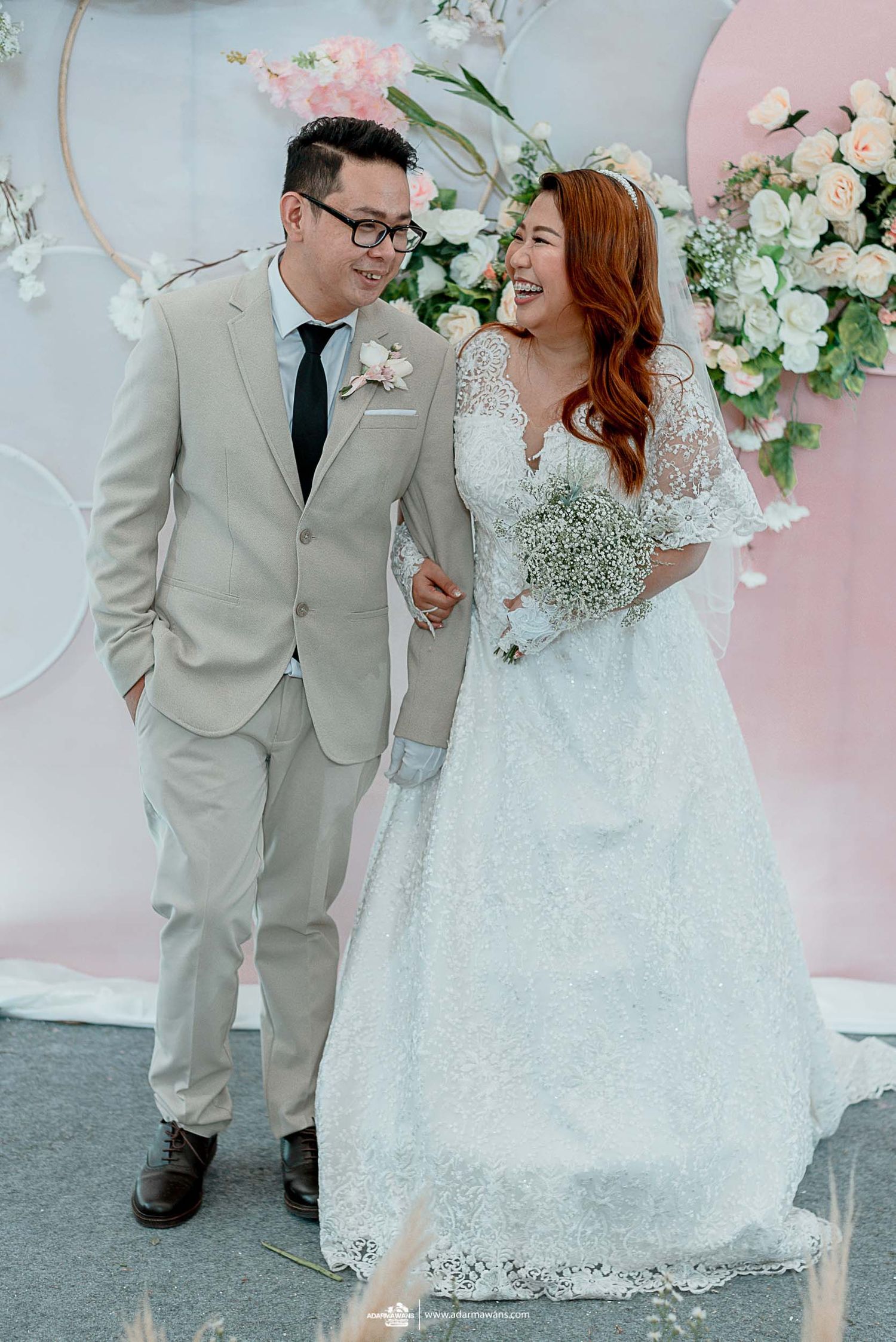 Happy couple sharing joyful moment against pink floral wedding backdrop.