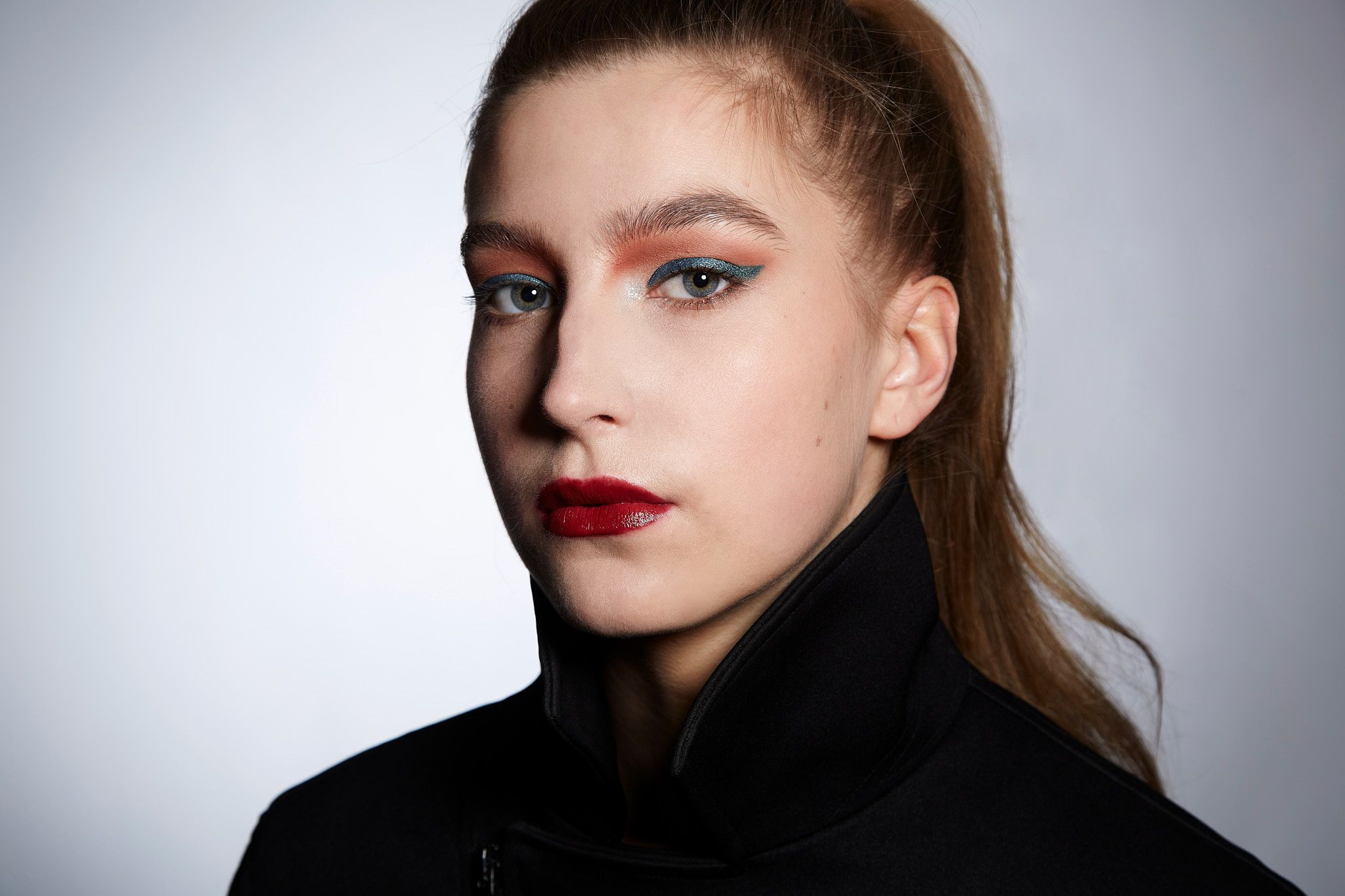 Dramatic portrait shot with dark turtleneck and striking makeup against grey background with bold expression.