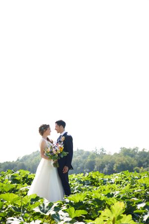 Newlyweds standing in field of large green leaves holding bright bouquet.