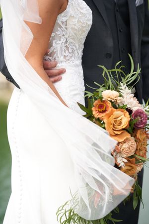 Close-up detail of white lace wedding dress with veil blowing in the wind and autumn-colored bouquet and grooms arm around her waiste.
