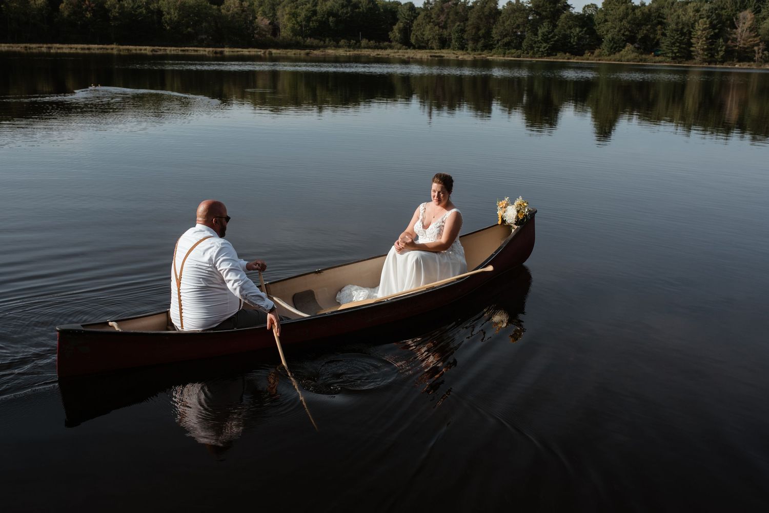 Newlyweds paddle in a wooden canoe across a serene lake during their wedding day.