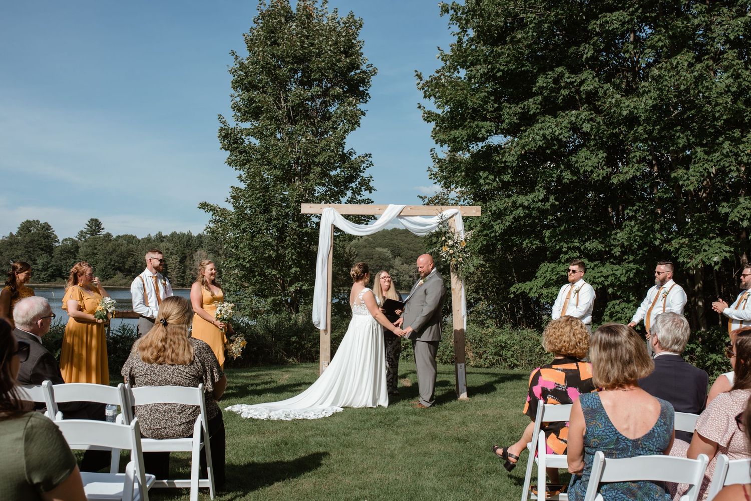 Outdoor wedding ceremony under wooden arch with white fabric draping and guests seated on lawn.