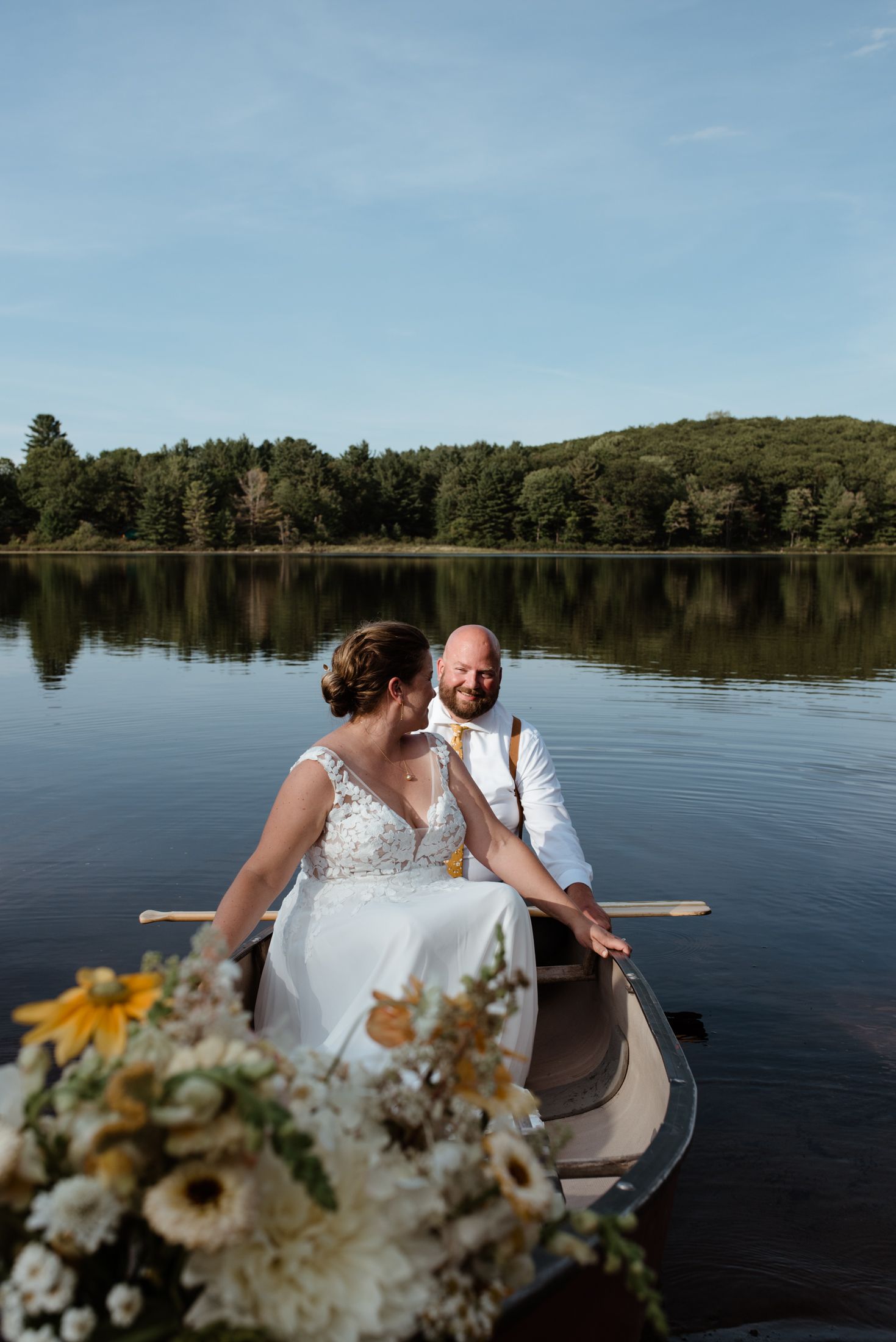 Couple sitting in a wooden boat on a serene lake surrounded by trees with a bouquet of flowers in the foreground.