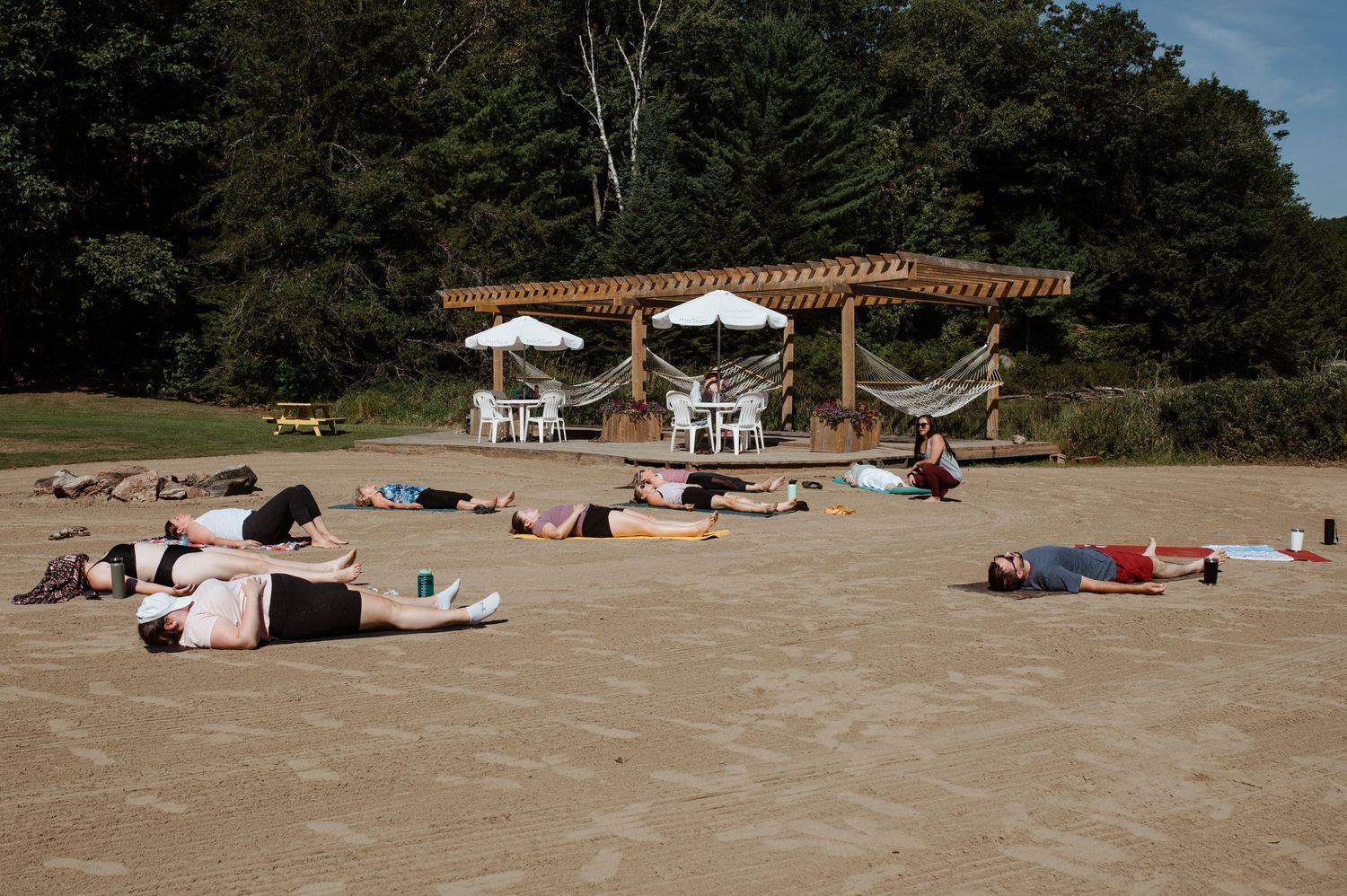 A group of people practicing yoga outdoors on mats under a wooden pergola on a sunny day.