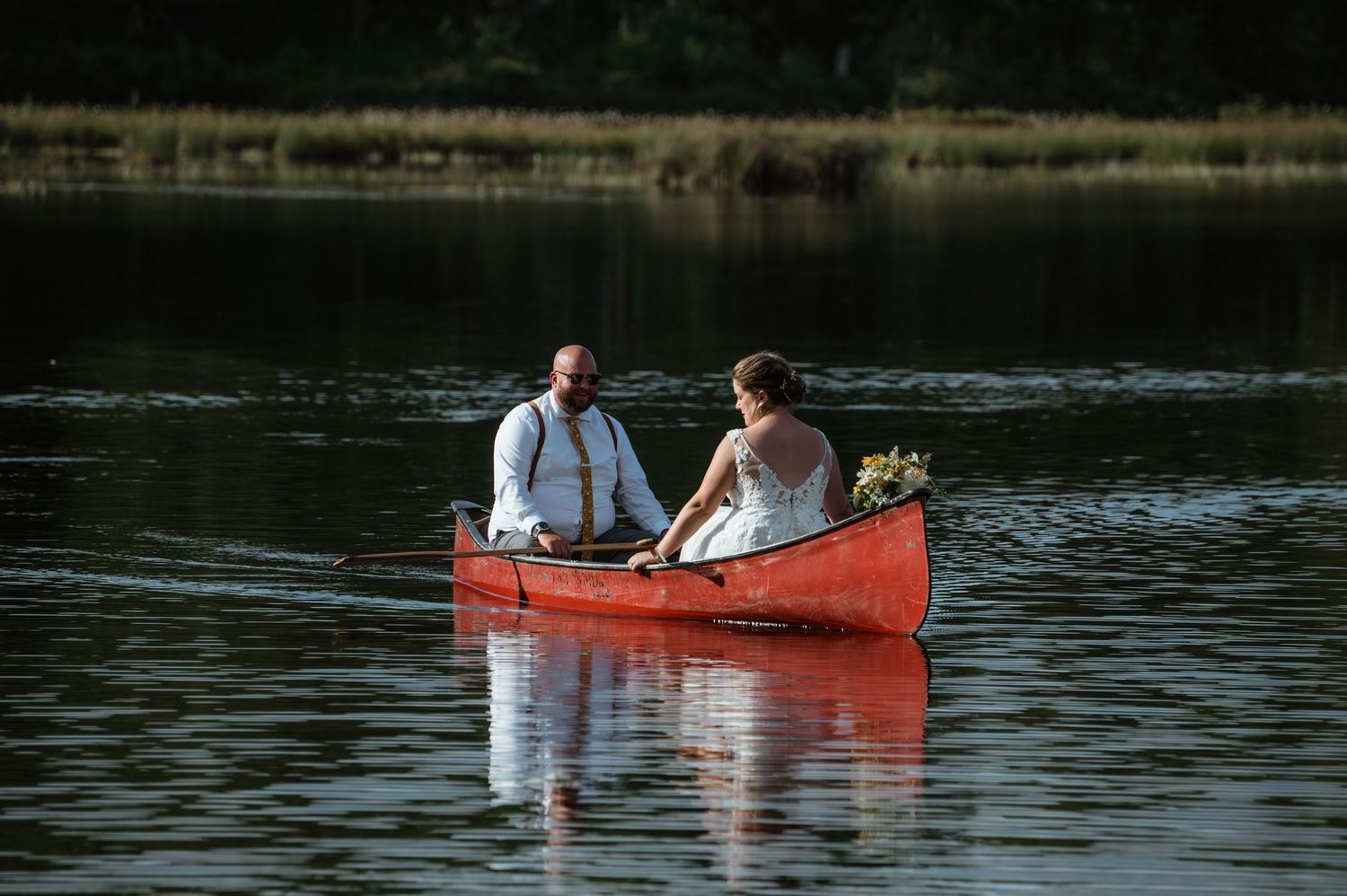 Couple in wedding attire rowing a red canoe on calm lake waters.