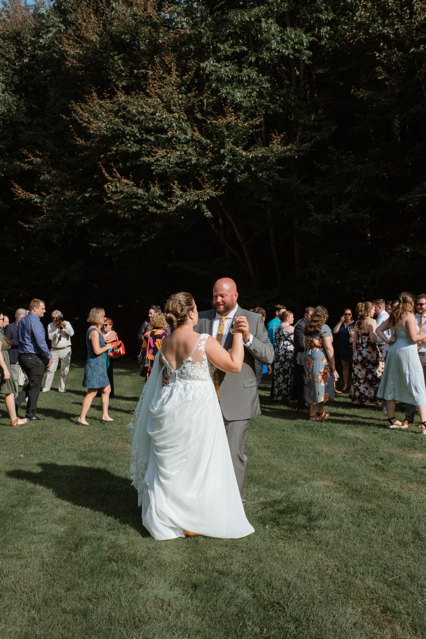 A couple shares their first dance on a sunny lawn surrounded by wedding guests in a garden setting.
