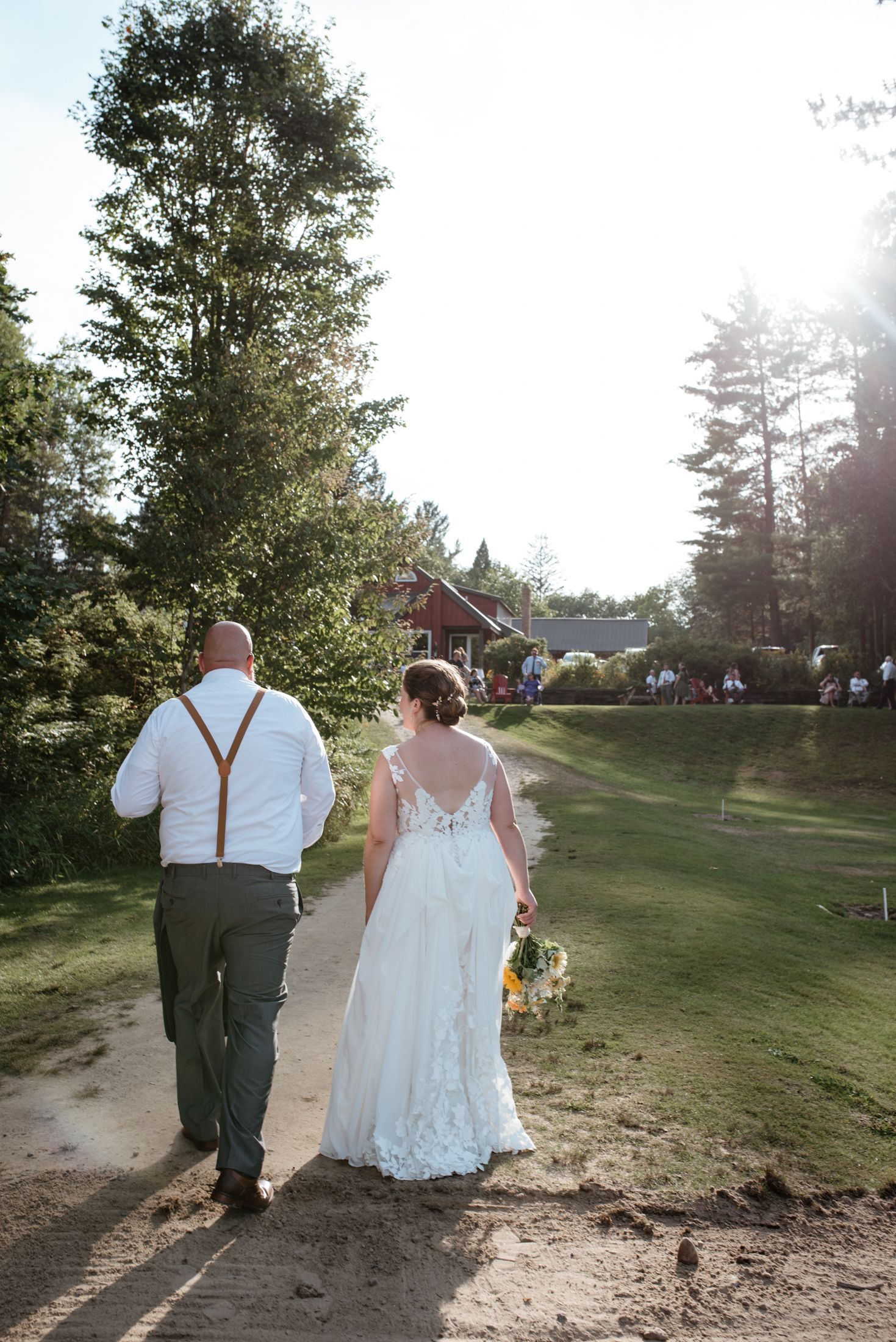 Couple walking down a dirt path towards a red barn during a sunny outdoor wedding celebration.