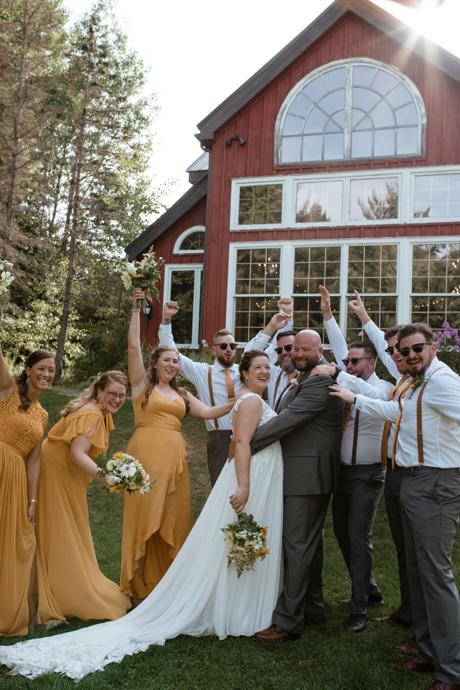 Wedding party poses playfully in front of a red barn-style venue with bridesmaids in mustard yellow dresses.