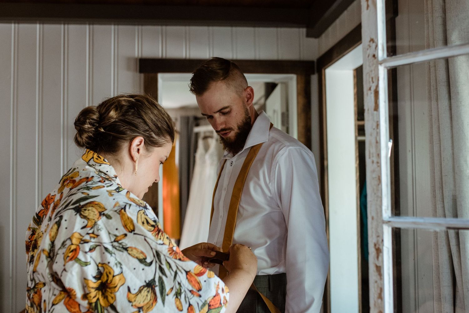 A floral-patterned shirt helps with buttoning suspenders on a white dress shirt in a rustic interior setting.
