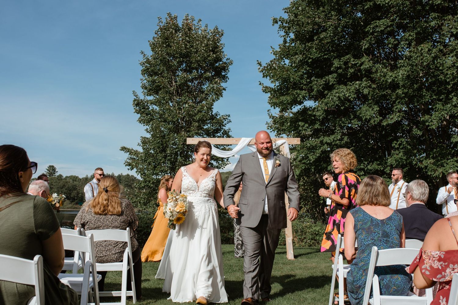 Wedding ceremony recessional on sunny day with guests seated on white chairs outdoors.
