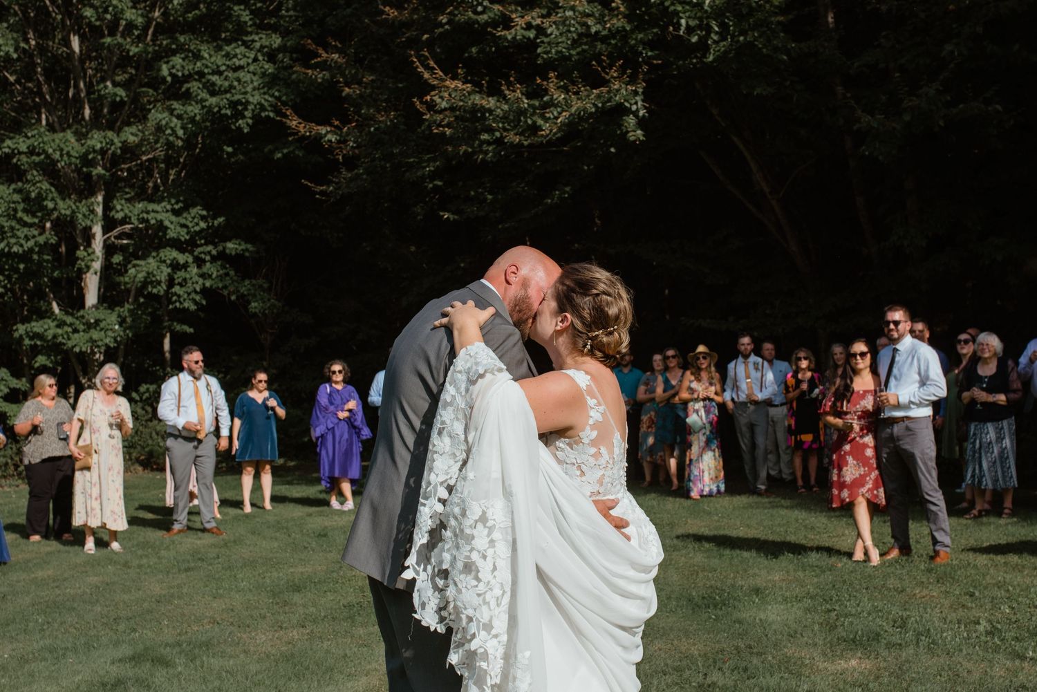 Wedding couple shares an intimate dance moment on a green lawn while guests watch from a distance.