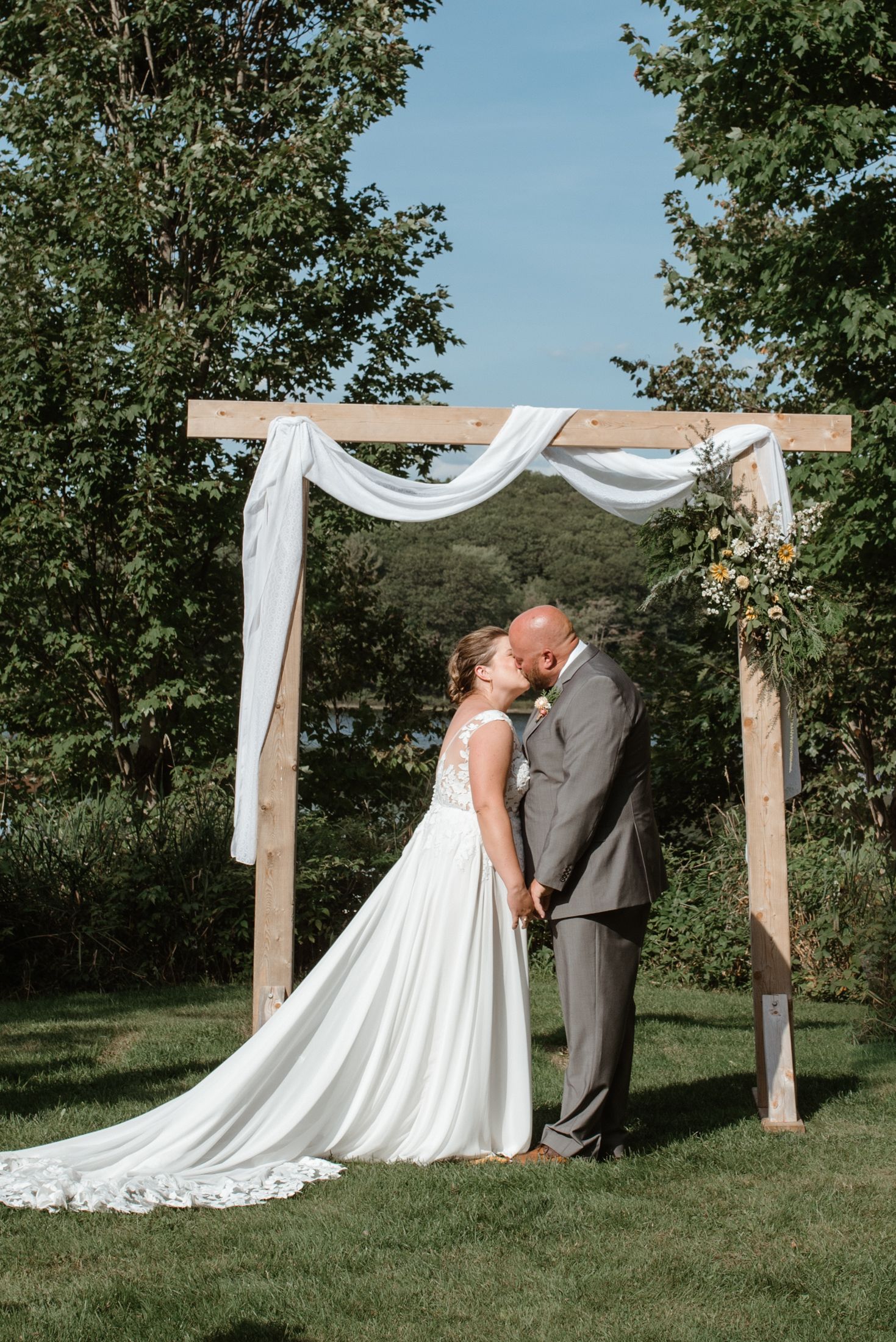 Wedding couple sharing intimate moment under wooden arch draped with white fabric against natural backdrop.