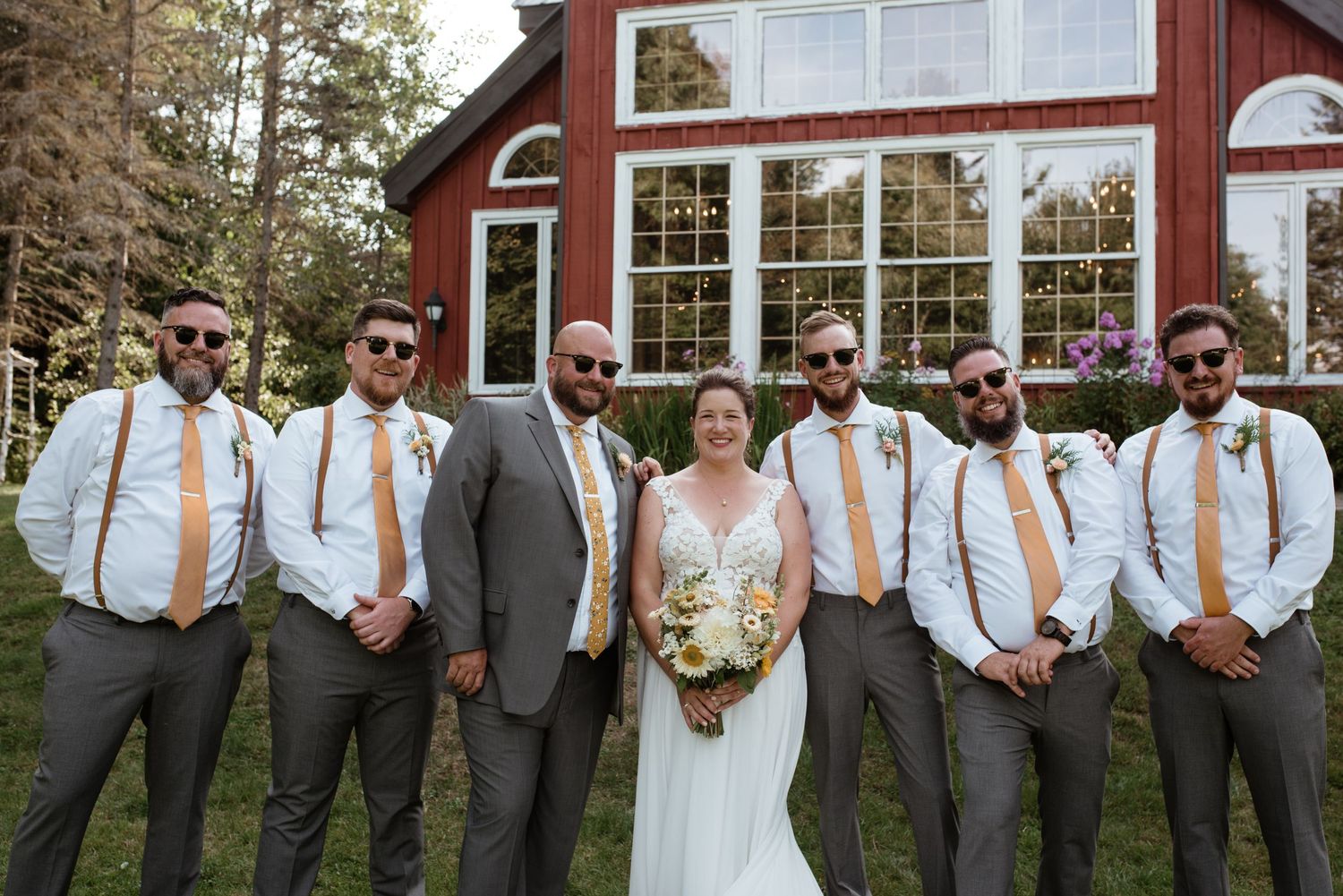Wedding party poses formally in front of red barn with groomsmen in suspenders and gray attire.