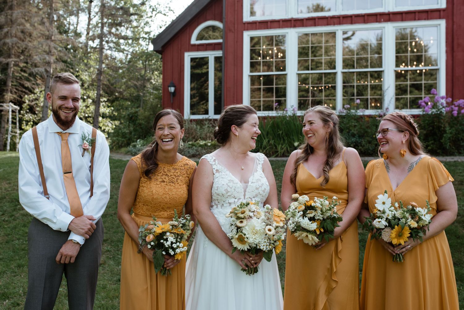 Wedding party poses in front of red barn venue with bridesmaids wearing golden yellow dresses.