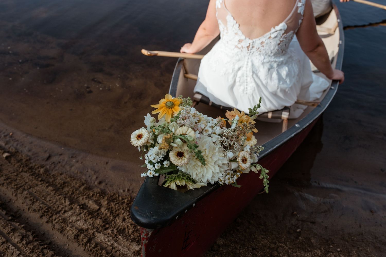 Wedding bouquet with white and yellow flowers rests in a red canoe on lake shore.