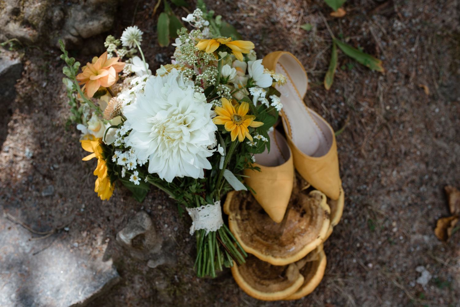 A bridal bouquet with white and yellow flowers next to yellow shoes on stone ground.