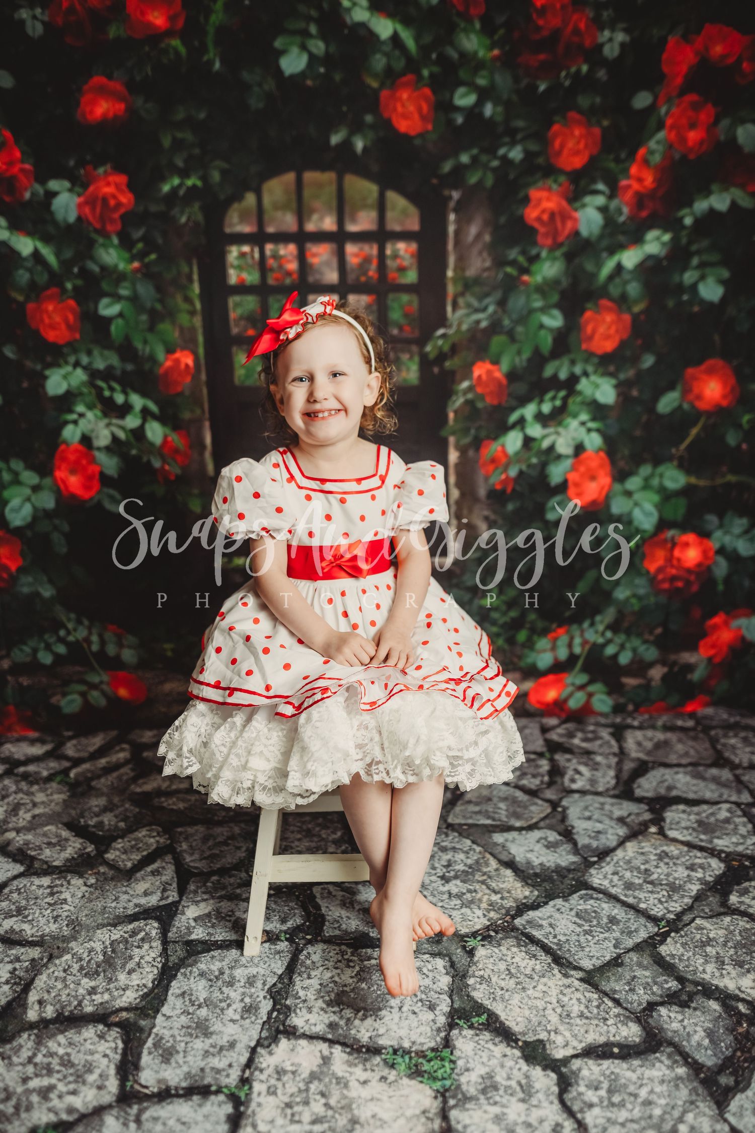 Child in polka dot dress sits on white chair against backdrop of climbing red roses.