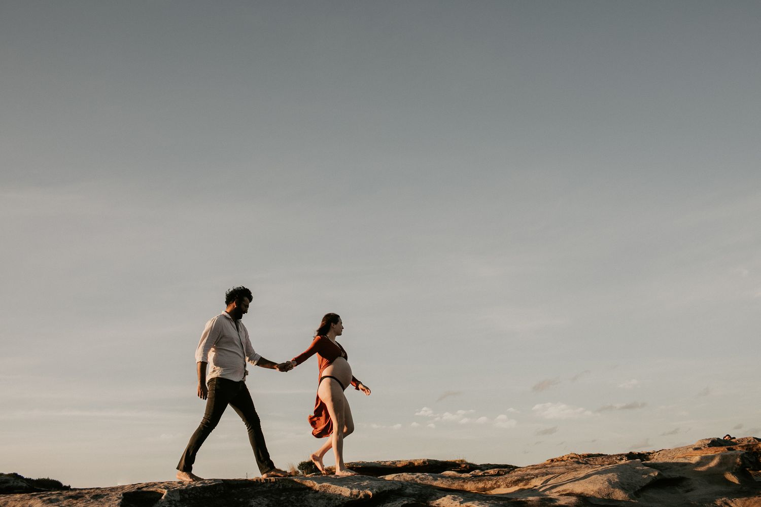A couple holds hands while walking across rocky terrain under a wide open sky at dusk.