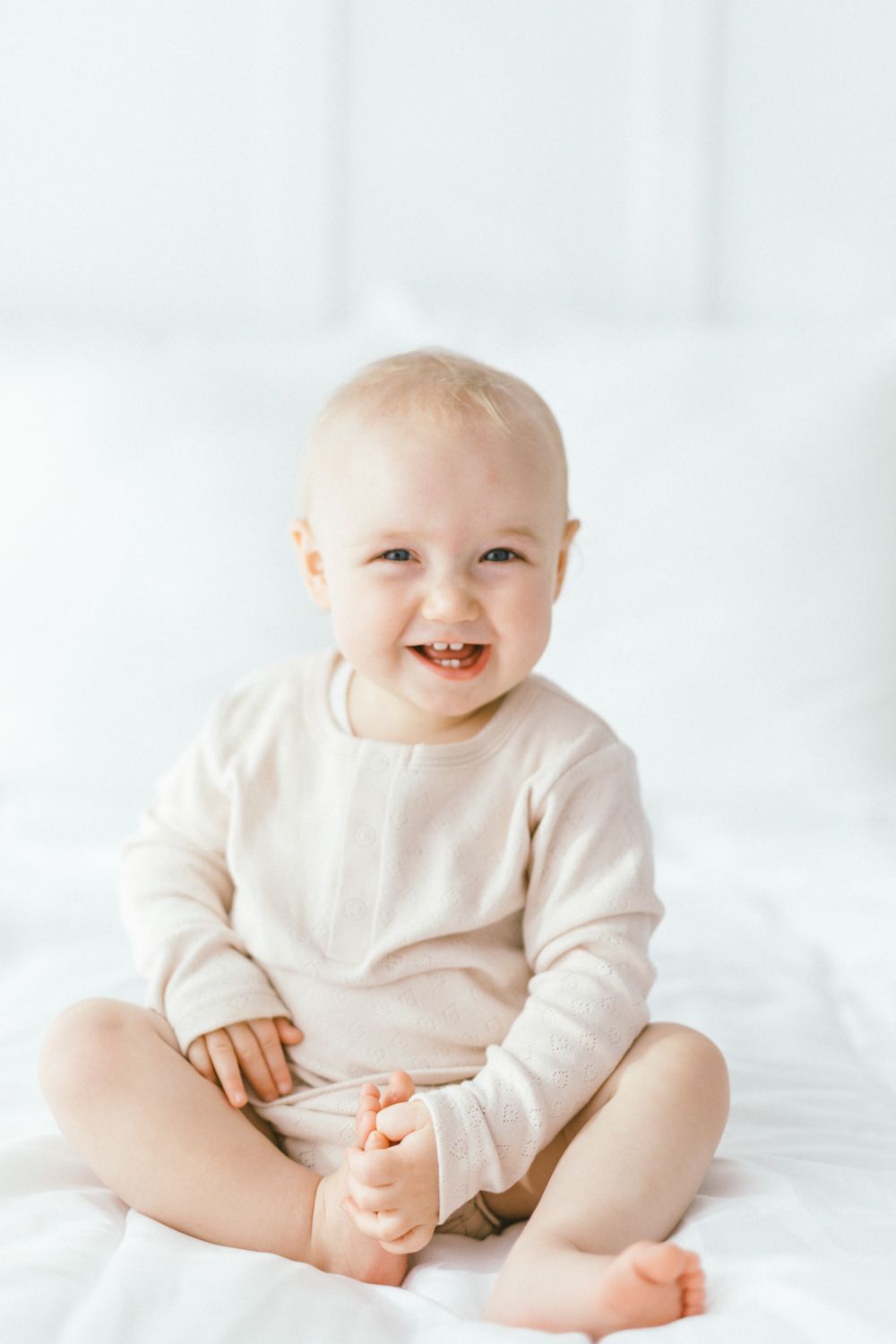Happy baby in cream colored outfit sitting on white bedding.