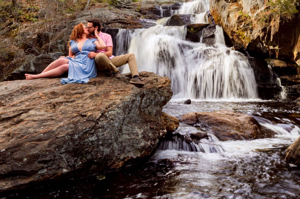 A couple in casual attire sits together on rocks beside a flowing waterfall.
