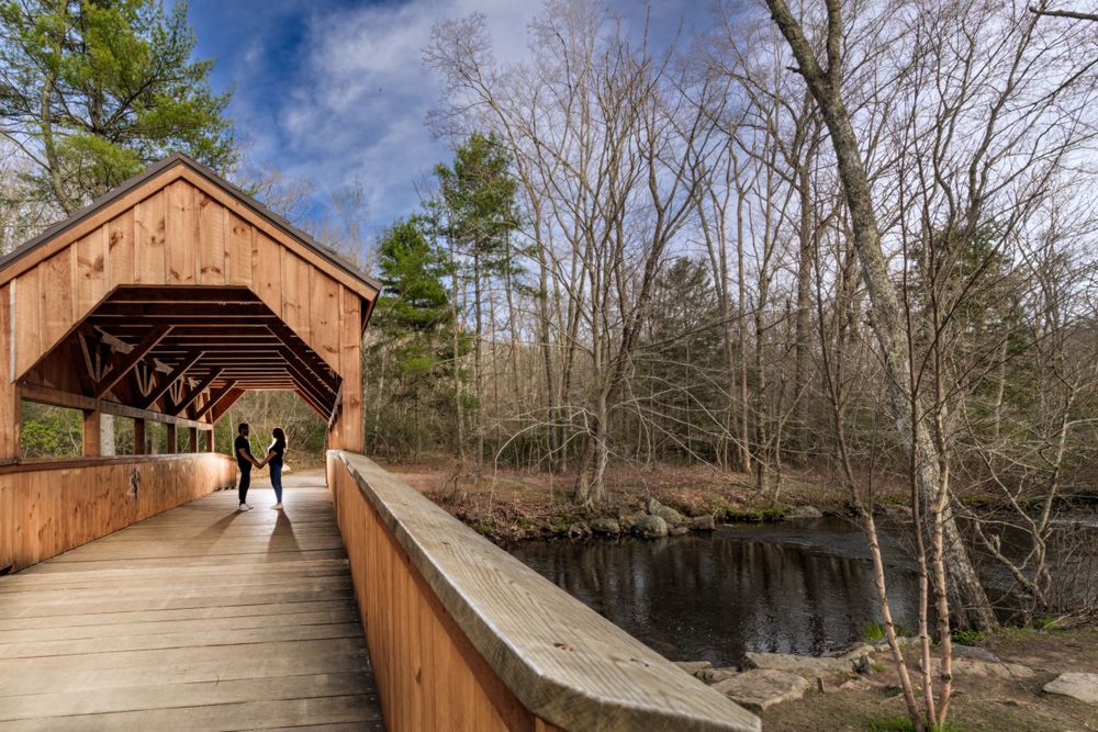A distant silhouette walks through a covered wooden bridge over water.