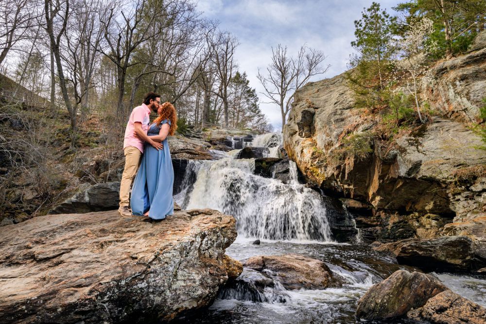Two figures share an intimate moment beside a scenic waterfall flowing over rugged rocks.