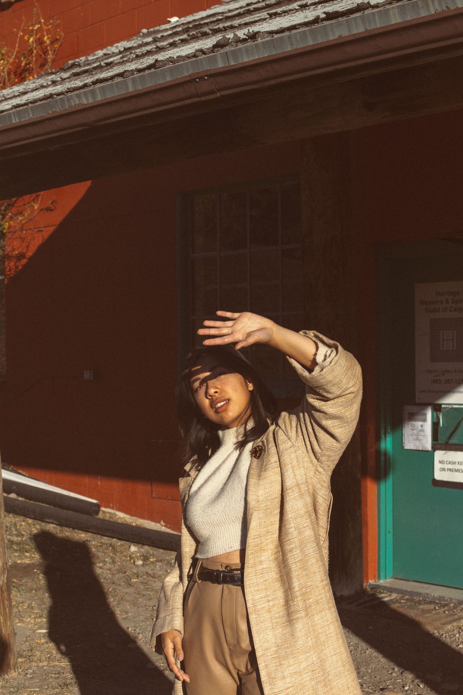 A stylish person in khaki pants and beige coat poses dramatically in front of a red building.