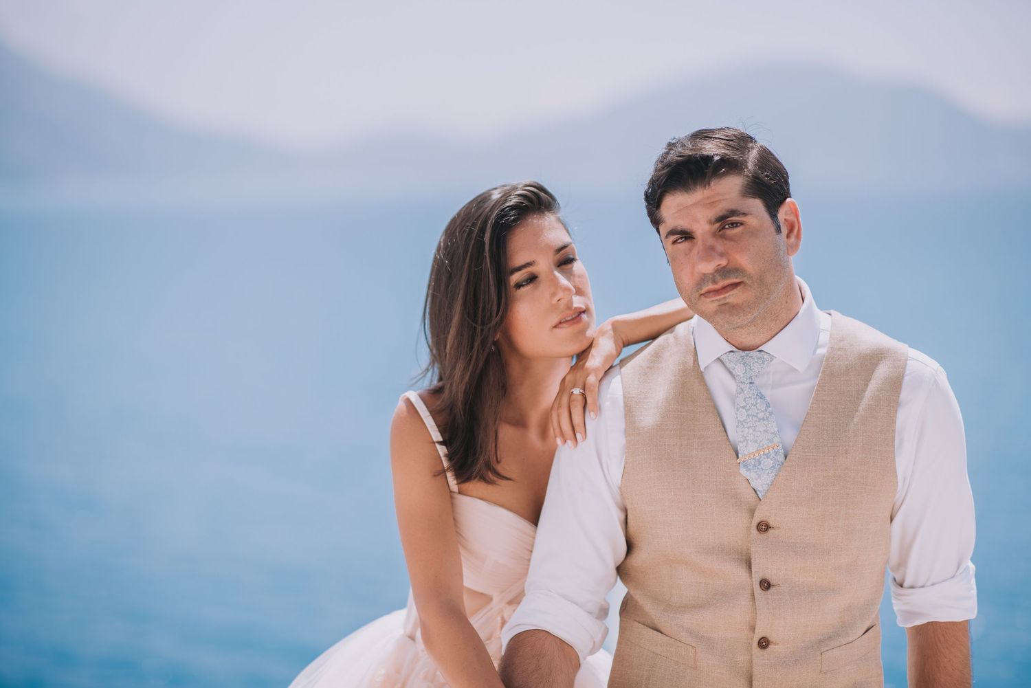 Romantic beachside photo session with couple in casual wedding attire against blue ocean backdrop.