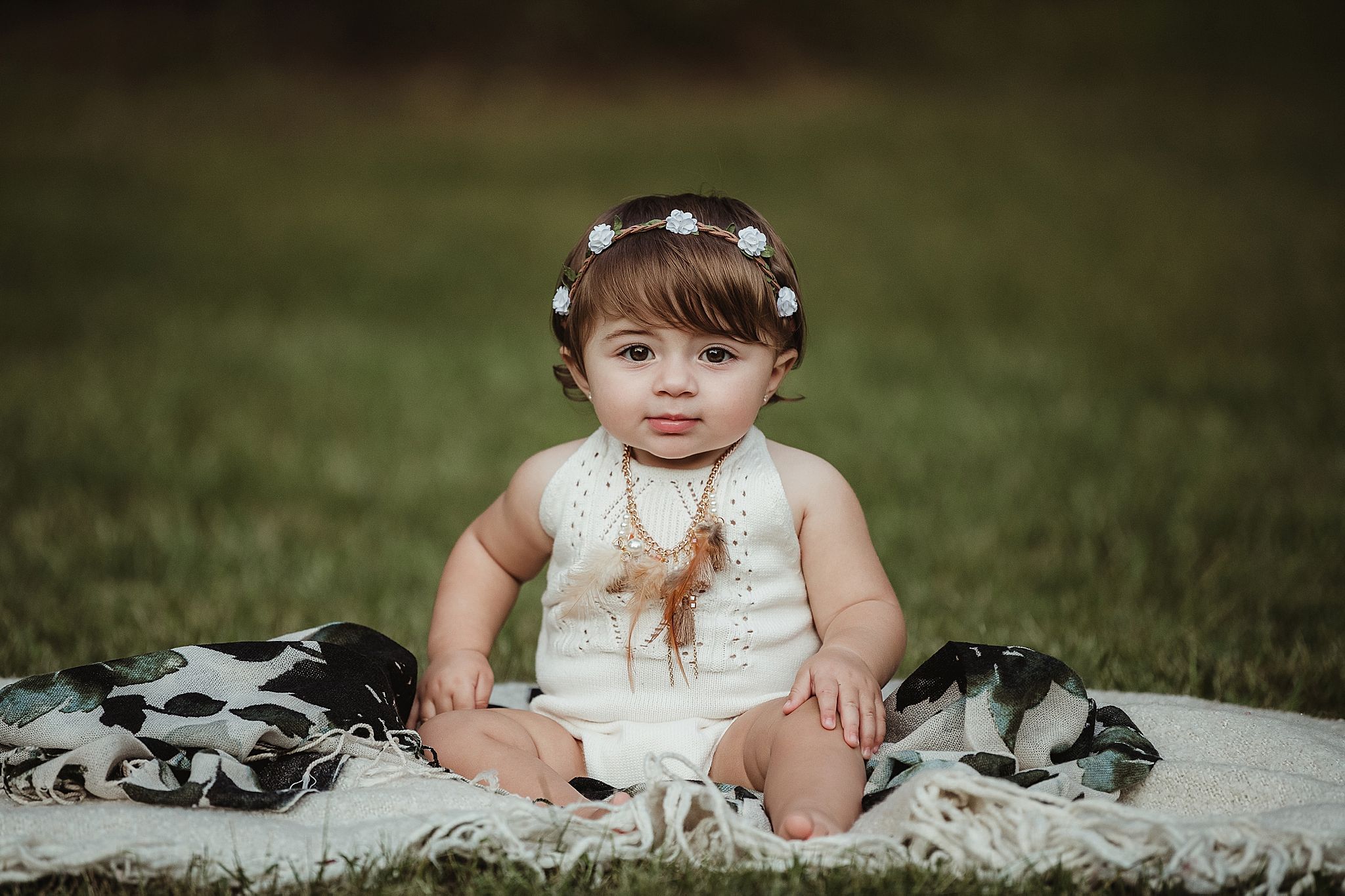 Baby in white dress sitting on blanket outdoors wearing floral crown headpiece.