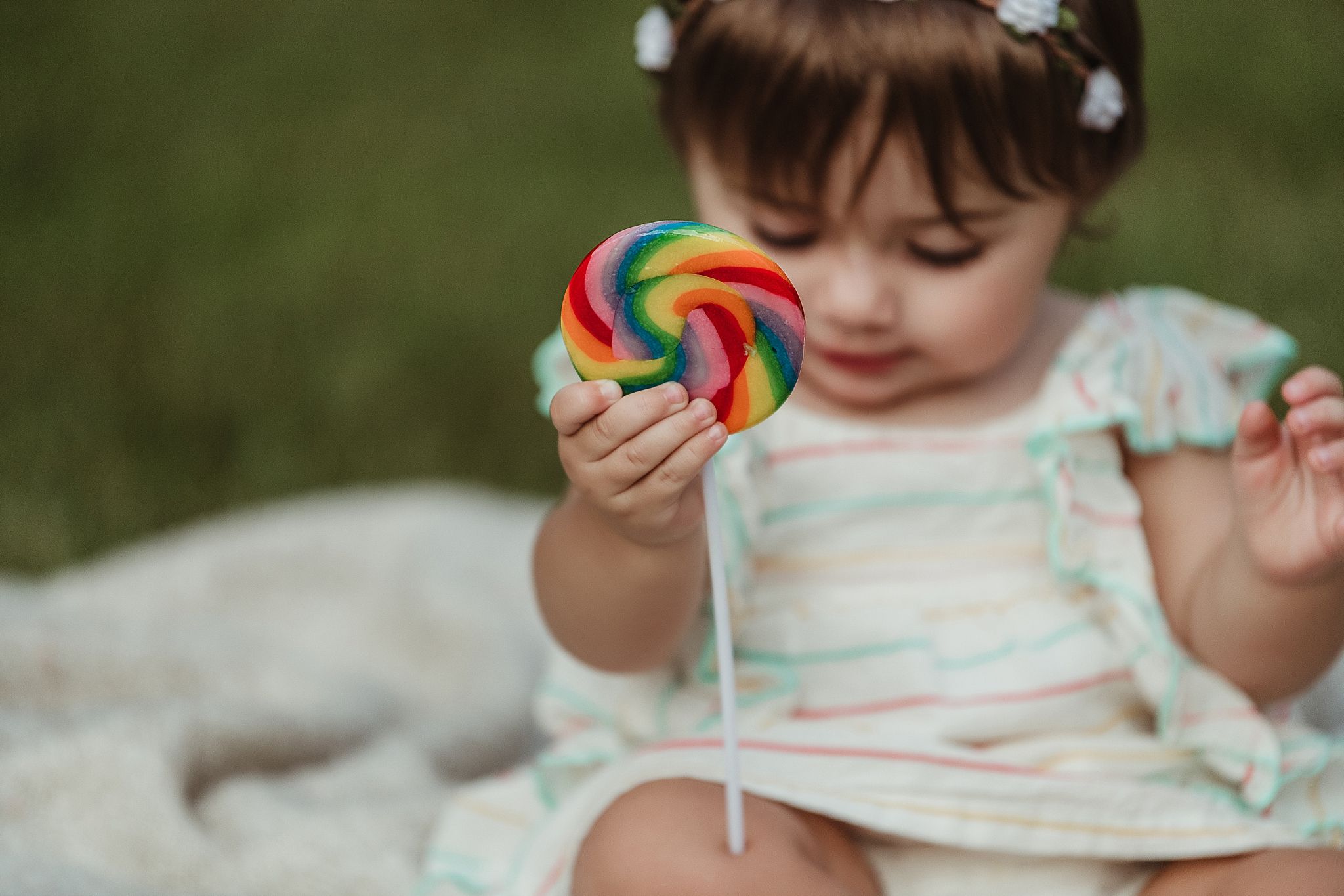 Child holding rainbow lollipop while sitting on white blanket outdoors.