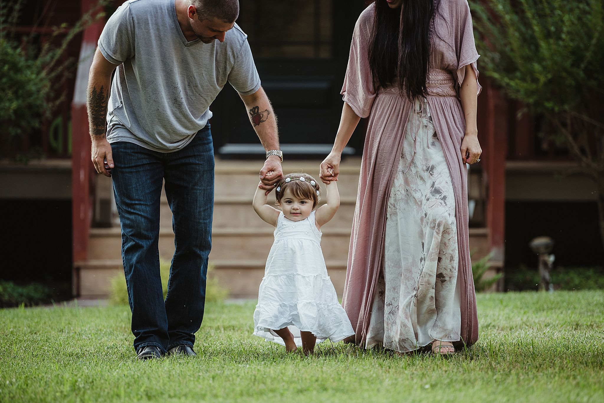 A family holds hands with a small child in a white dress while walking in a garden.