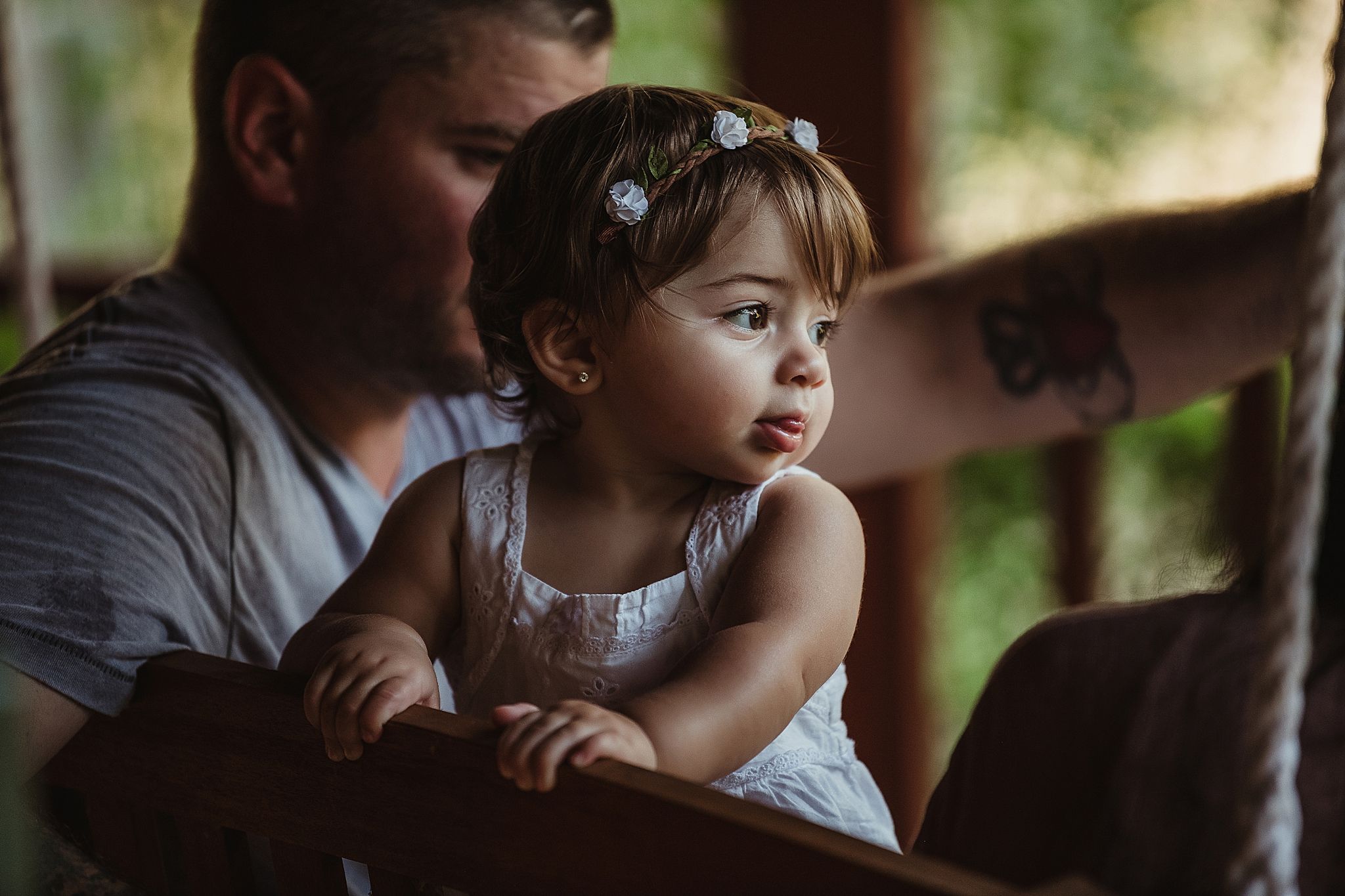 A small child in a white dress sits with an adult on a porch swing in soft natural lighting.