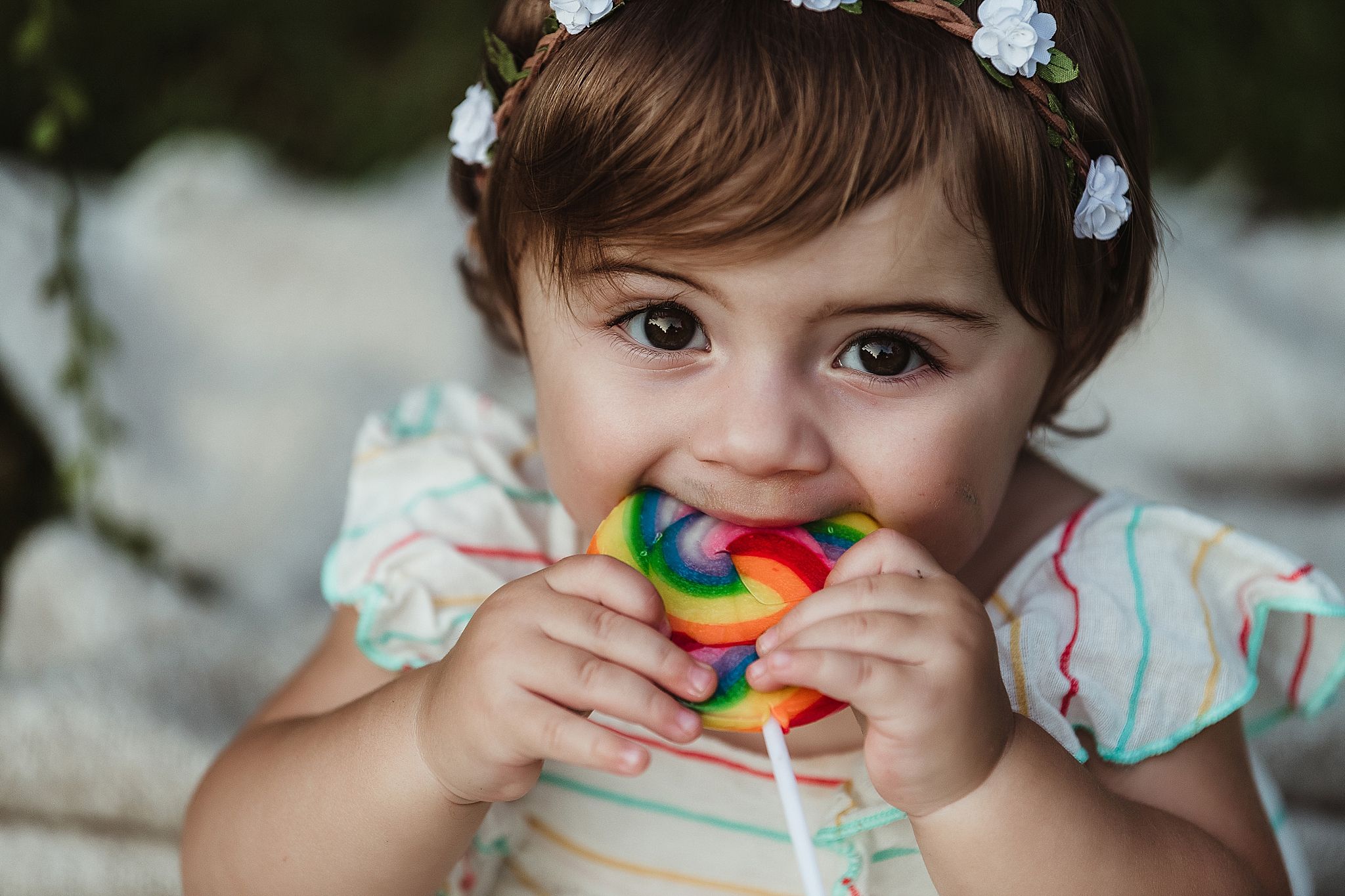 Child enjoying a rainbow swirl lollipop while wearing floral dress and flower crown.