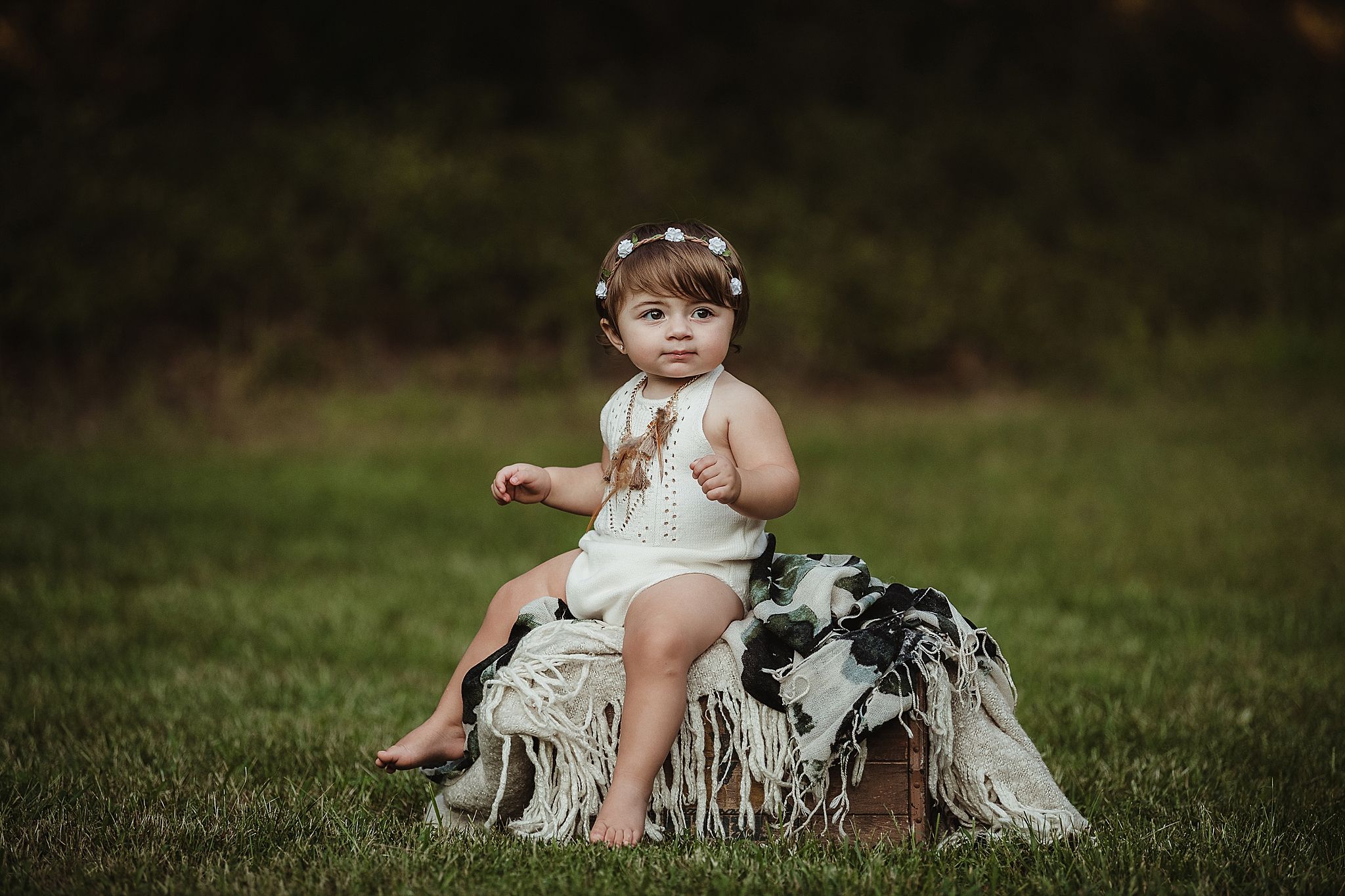 Baby in white romper sitting on textured blanket in grassy outdoor setting.