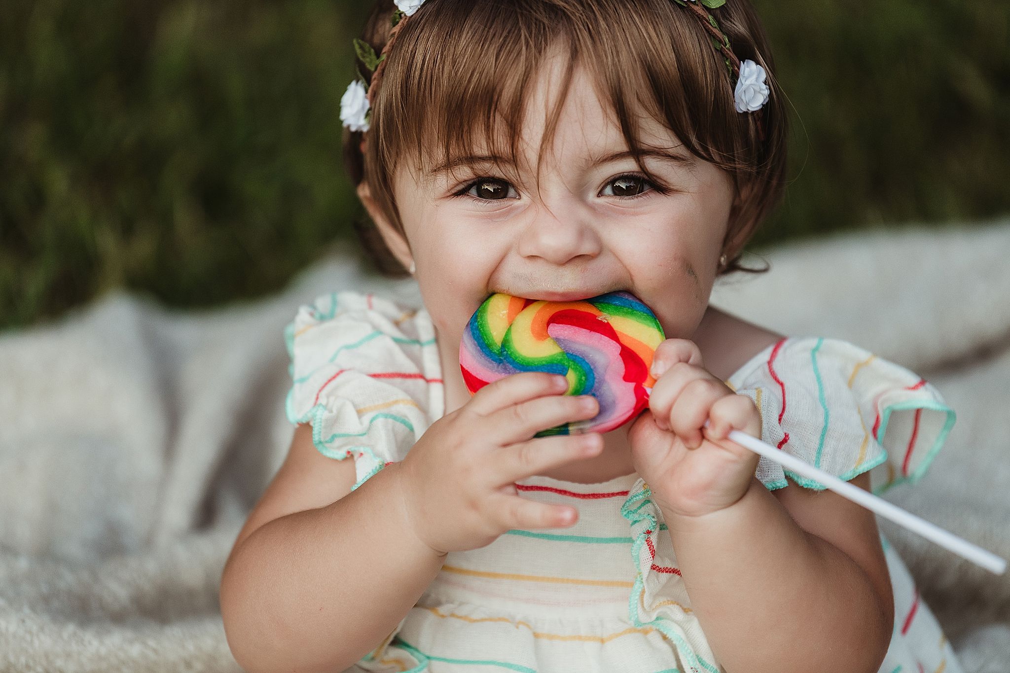 Small child in floral dress holding colorful lollipop while sitting.