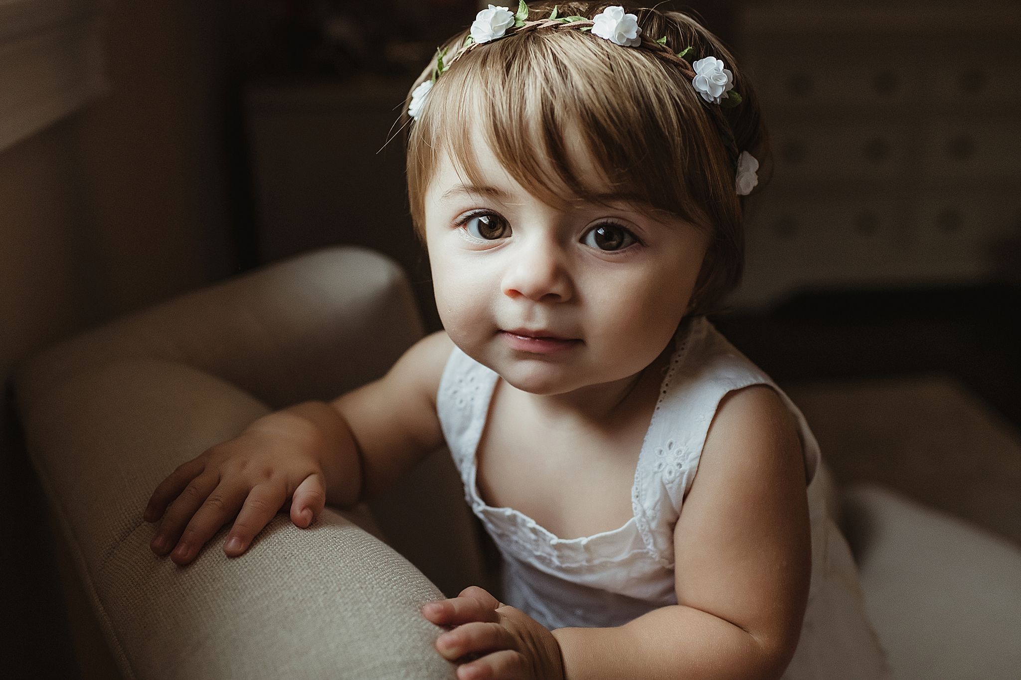 Close up of toddler in white dress and floral crown sitting in cozy indoor setting.