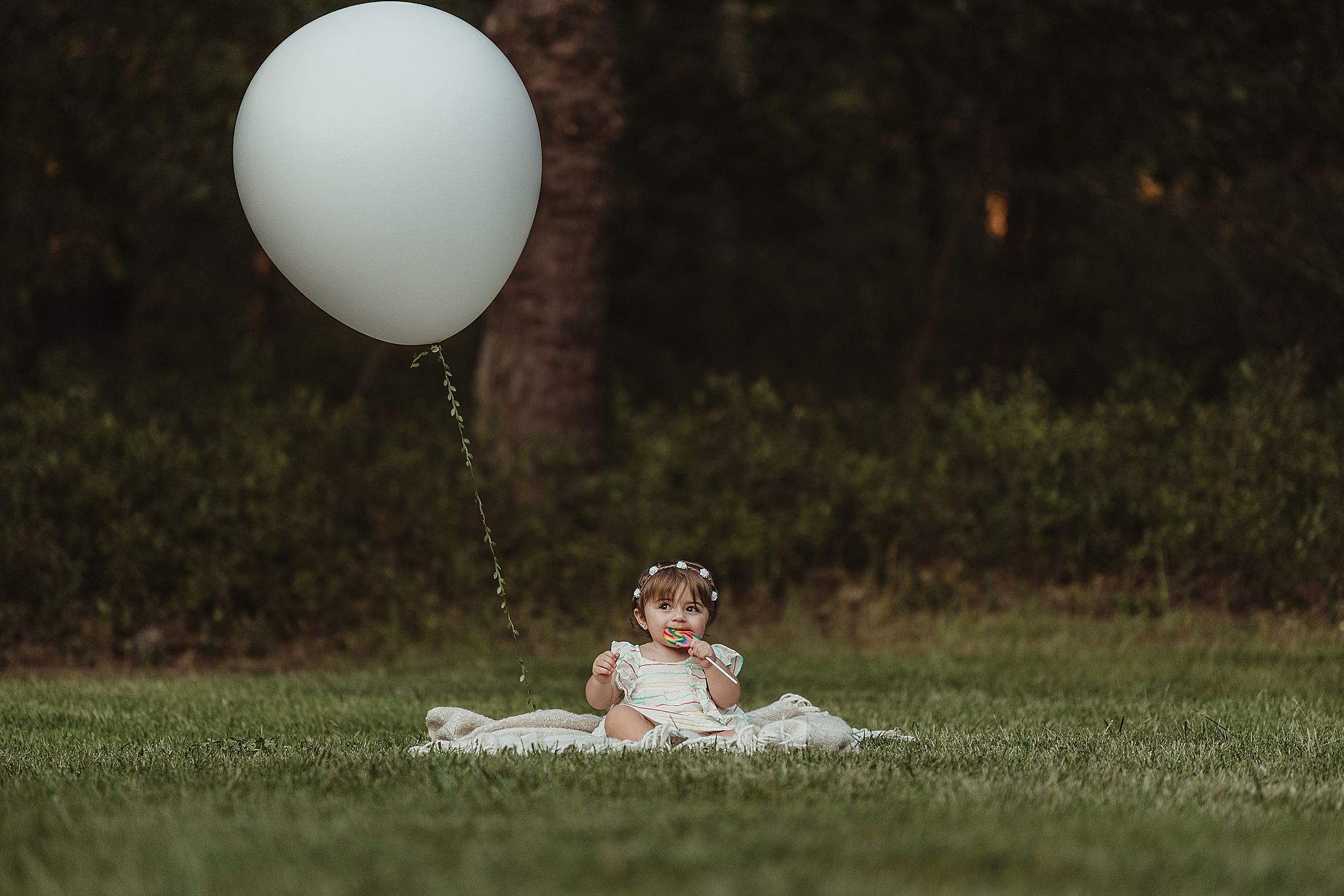 Child in white dress sitting on blanket with large white balloon in garden setting.