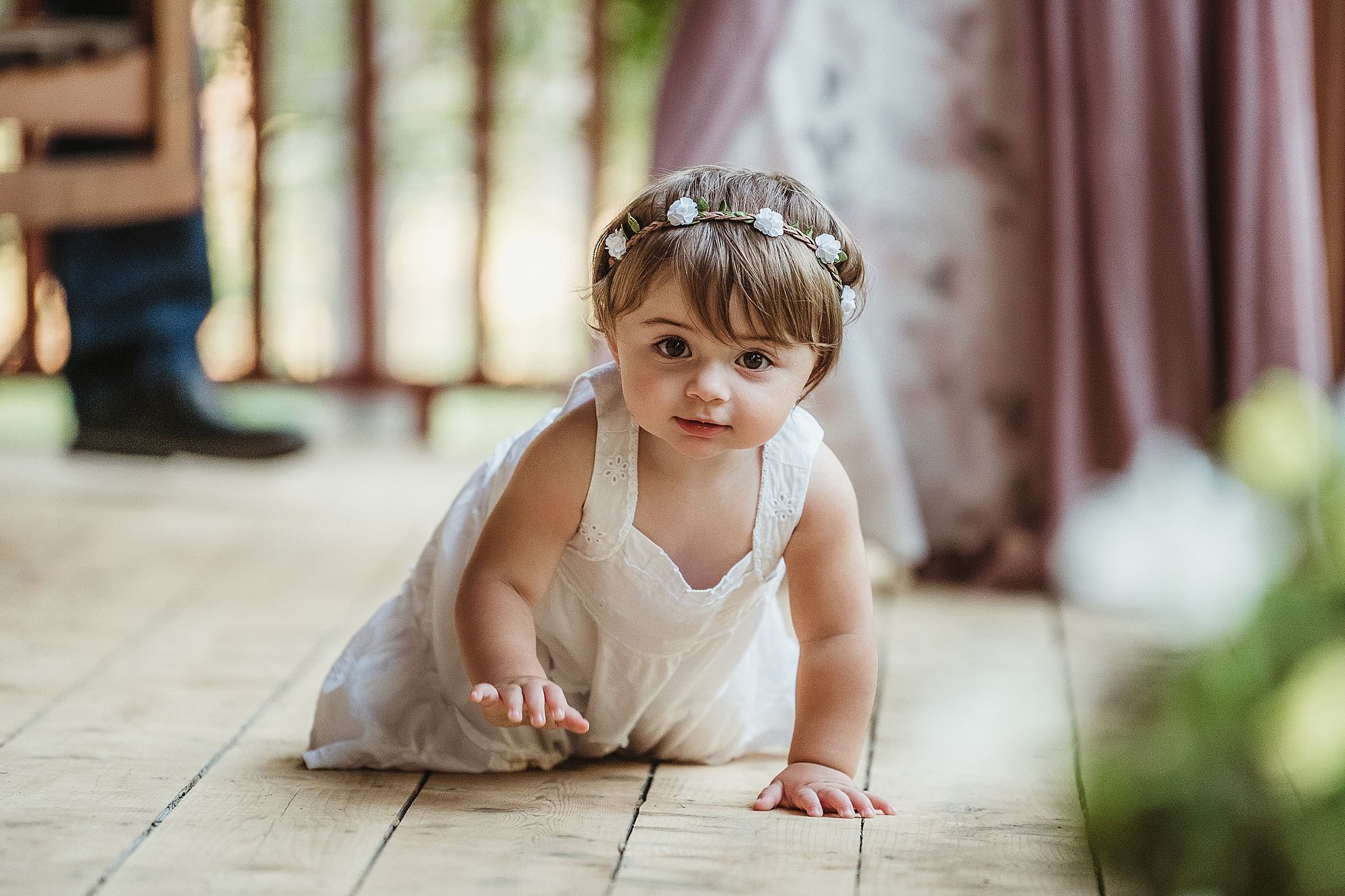 A small child in a white dress crawls on a wooden floor with natural light streaming in.