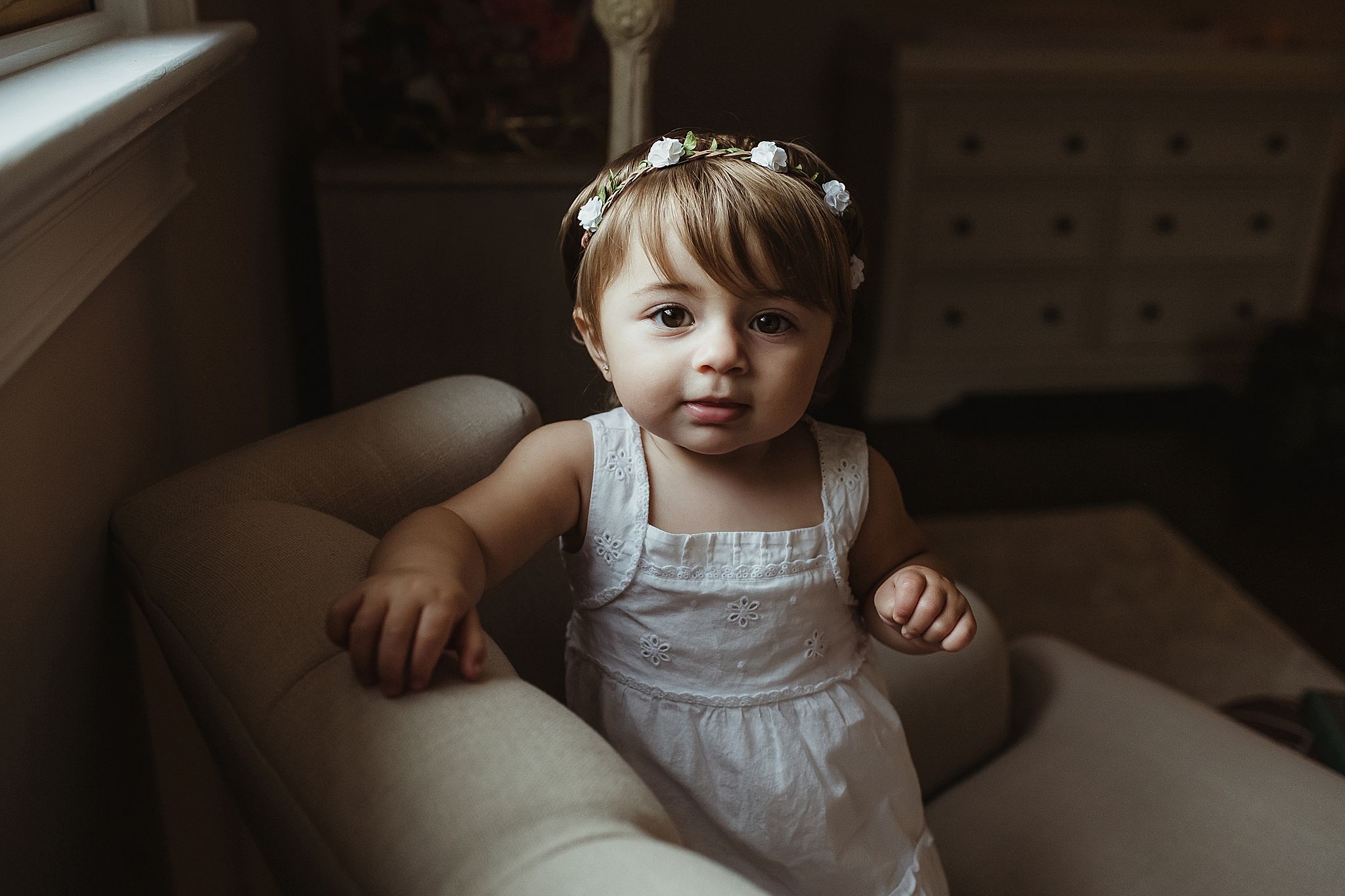 Indoor portrait of toddler in white dress sitting by window with floral crown.