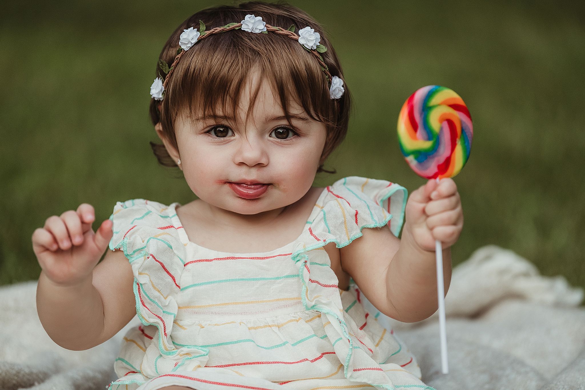 Child in white dress holds up colorful lollipop while sitting on blanket outdoors.