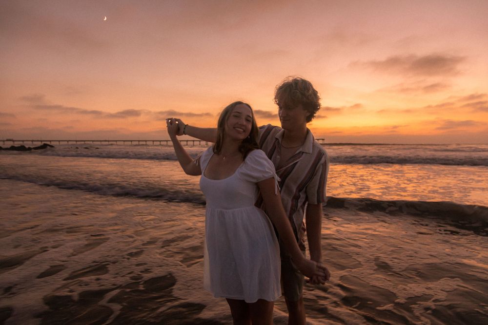 Romantic couple embraces on beach during vibrant sunset with pier visible in distance.