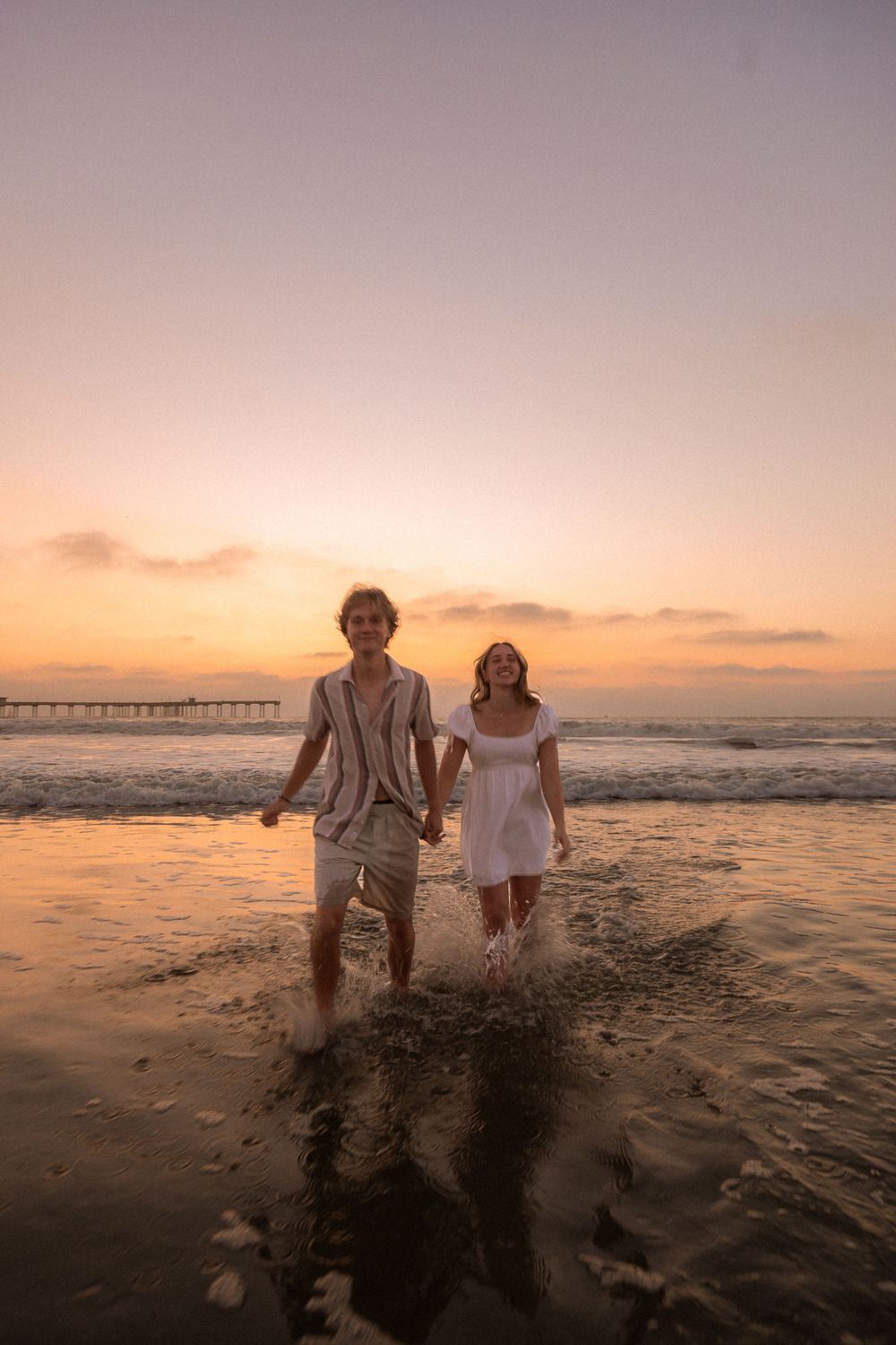 Couple holding hands while walking in the ocean waves during a golden sunset by a pier.