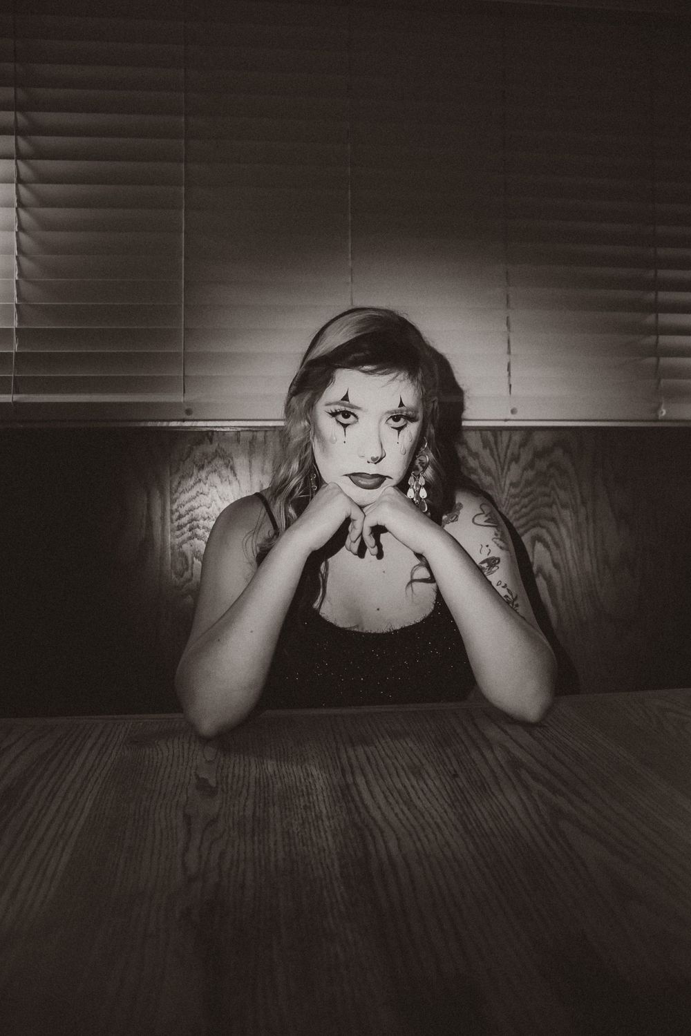 Dramatic black and white portrait with theatrical makeup at dimly lit table.