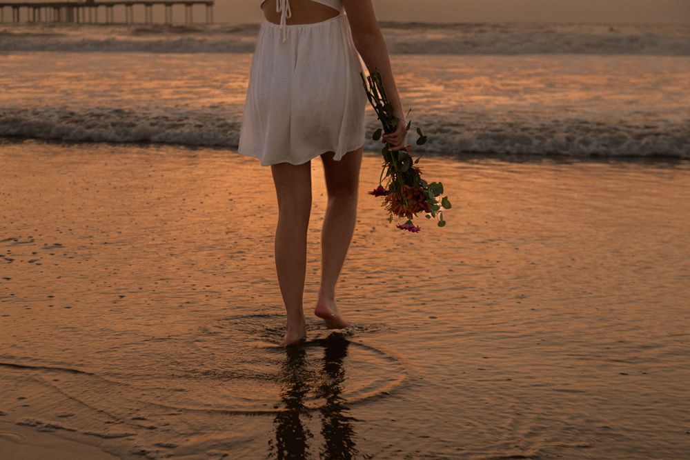 Close-up of legs walking in shallow water at sunset holding flowers on beach.