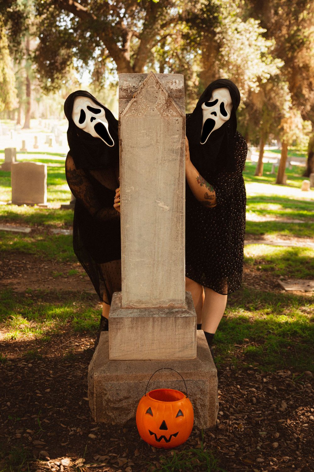 Two figures in Ghostface masks flank a cemetery headstone with an orange pumpkin decoration.