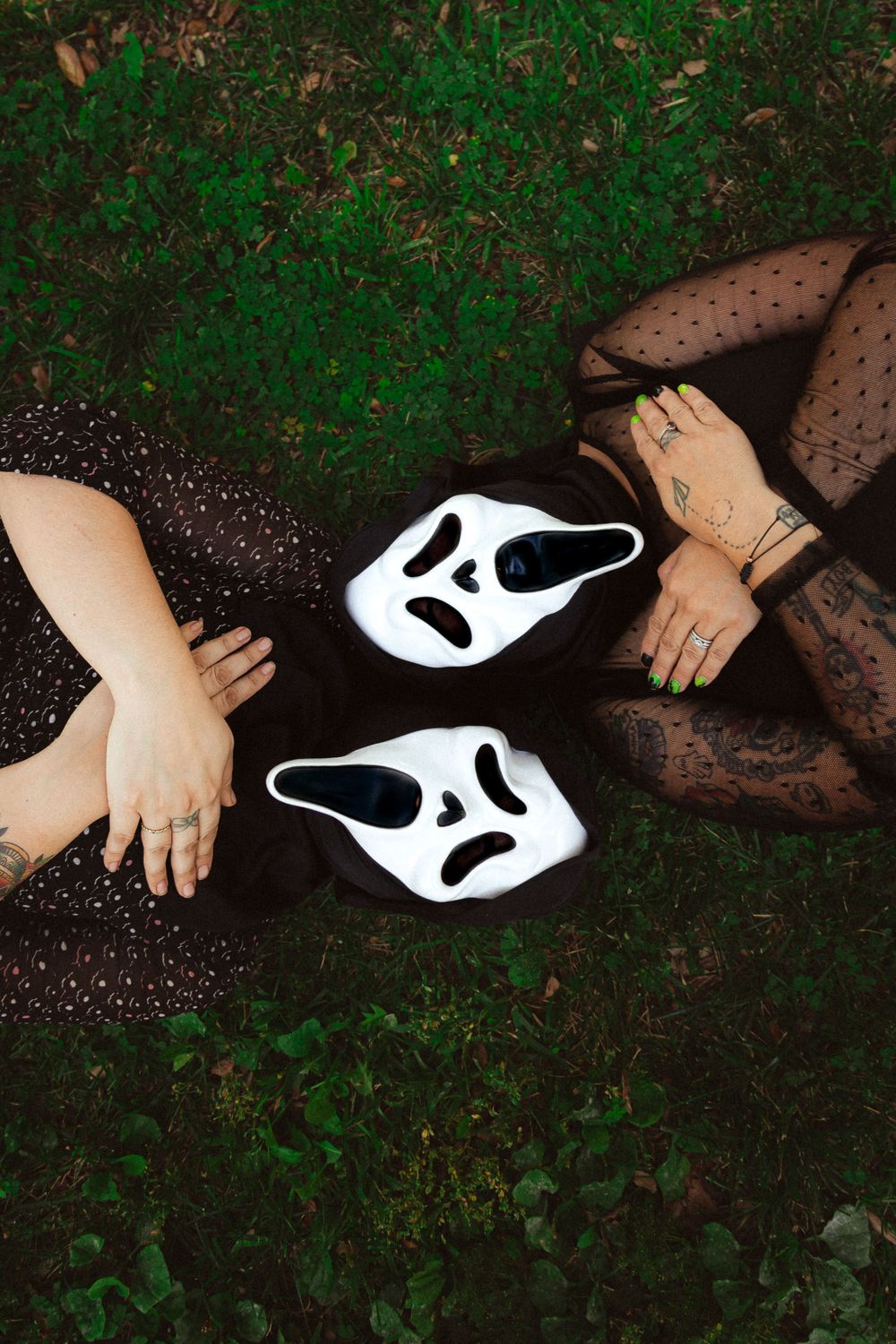 Two Scream-style masks on grass between hands wearing sheer black sleeves.