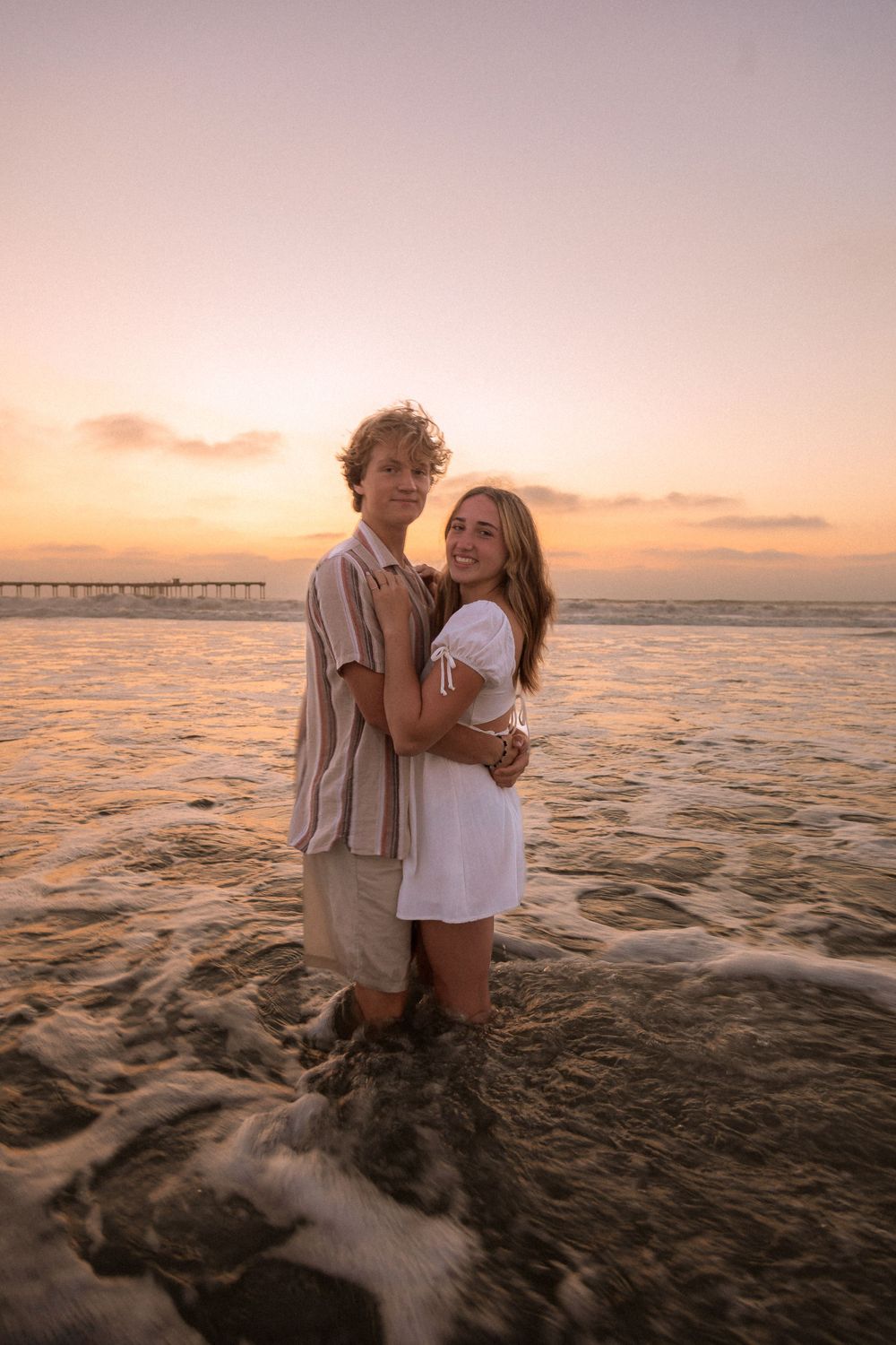 Couple embracing in ocean waves during colorful beach sunset.