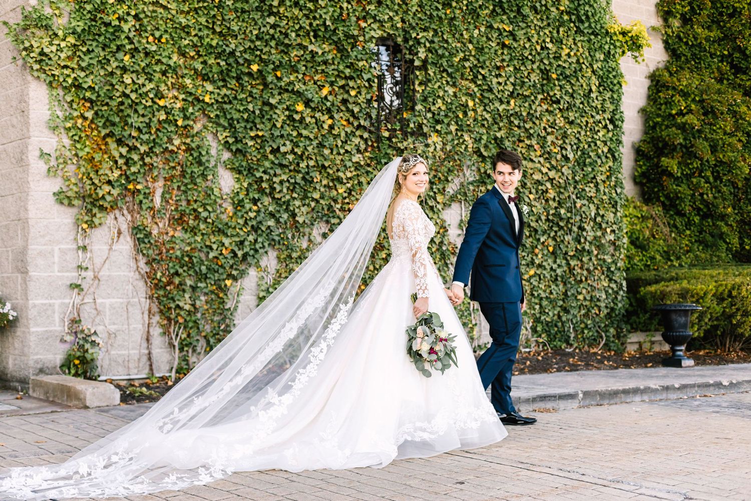 A bride with flowing veil walks with groom against an ivy-covered wall.