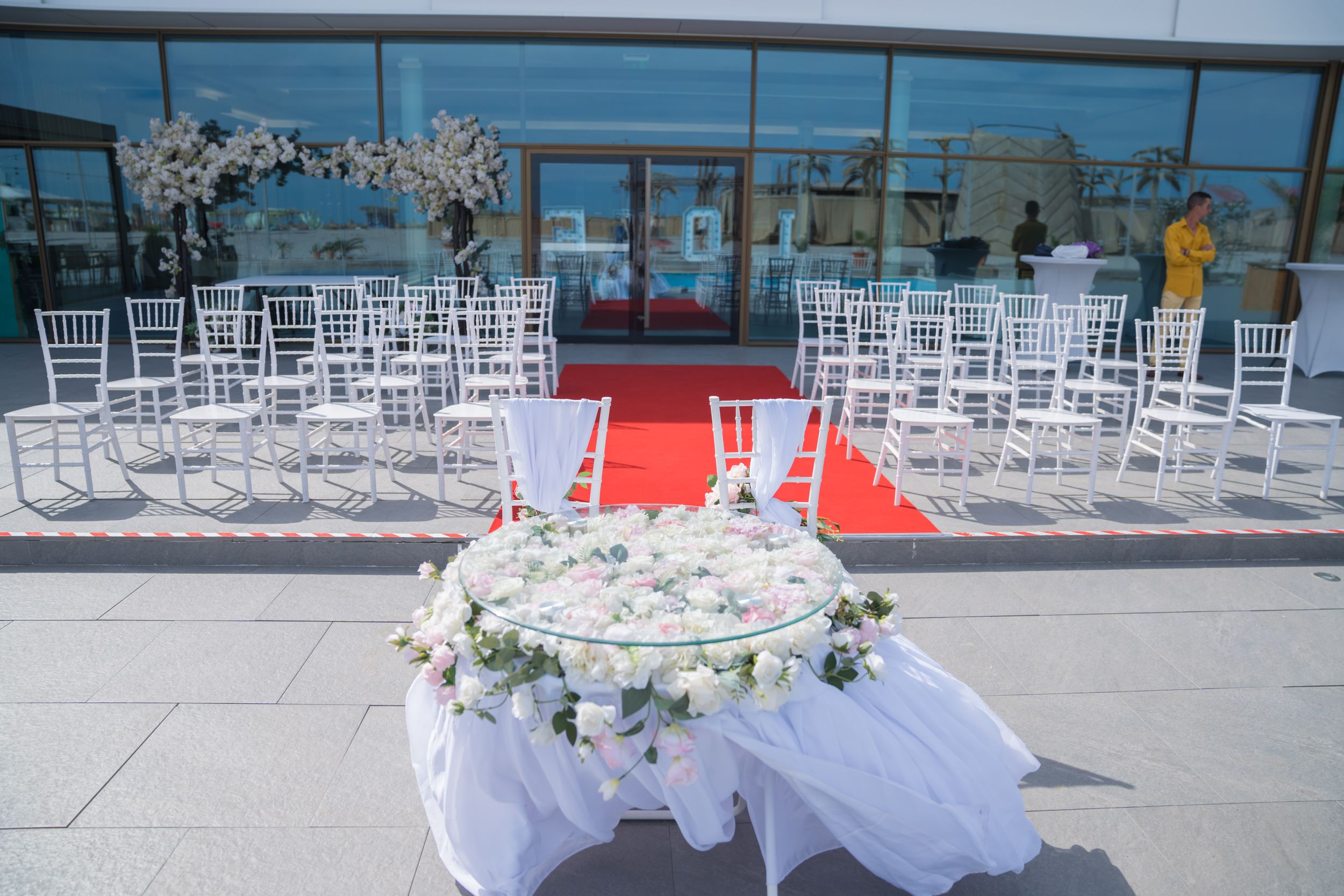 White ceremony chairs arranged outdoors with floral decorations and red carpet aisle.